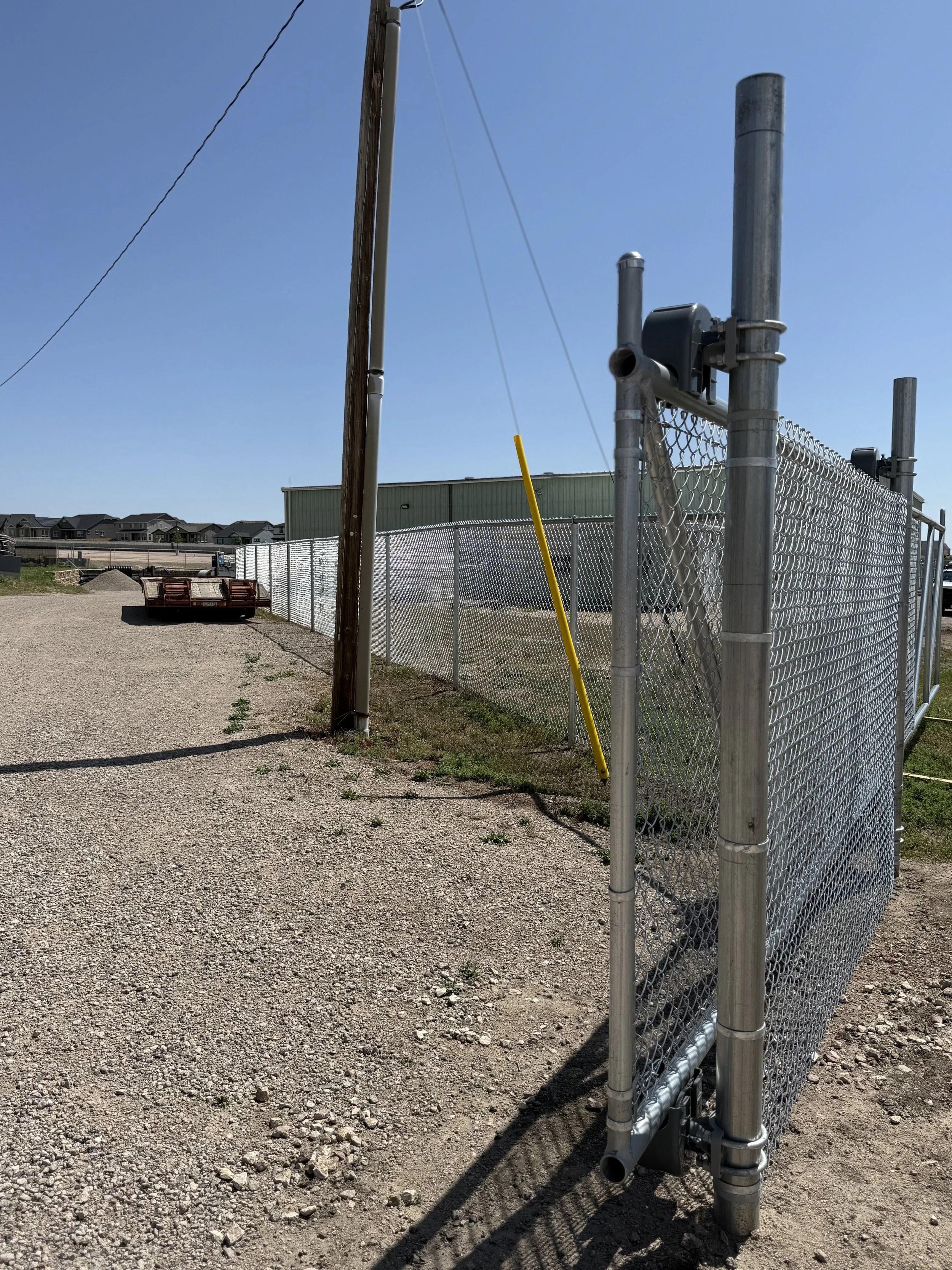 A gravel driveway with a chain-link fence on the right side, a utility pole leaning slightly, and a yellow pole leaning against the fence. In the distance, there is a green building and a red trailer on the gravel. Residential houses are visible beyo