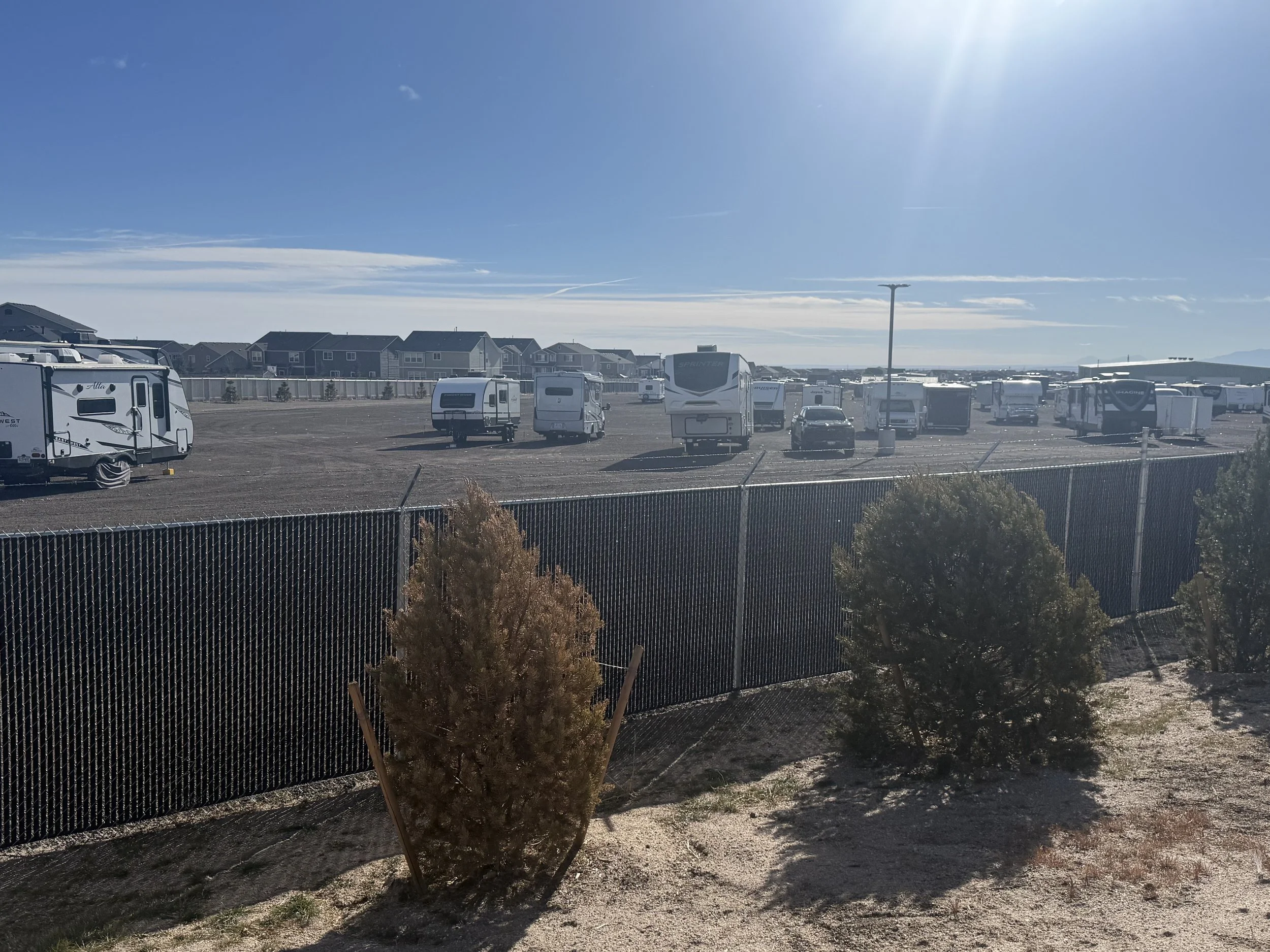 RVs and trailers parked in a large outdoor lot, with a chain-link fence in the foreground, under a clear blue sky with the sun shining brightly.