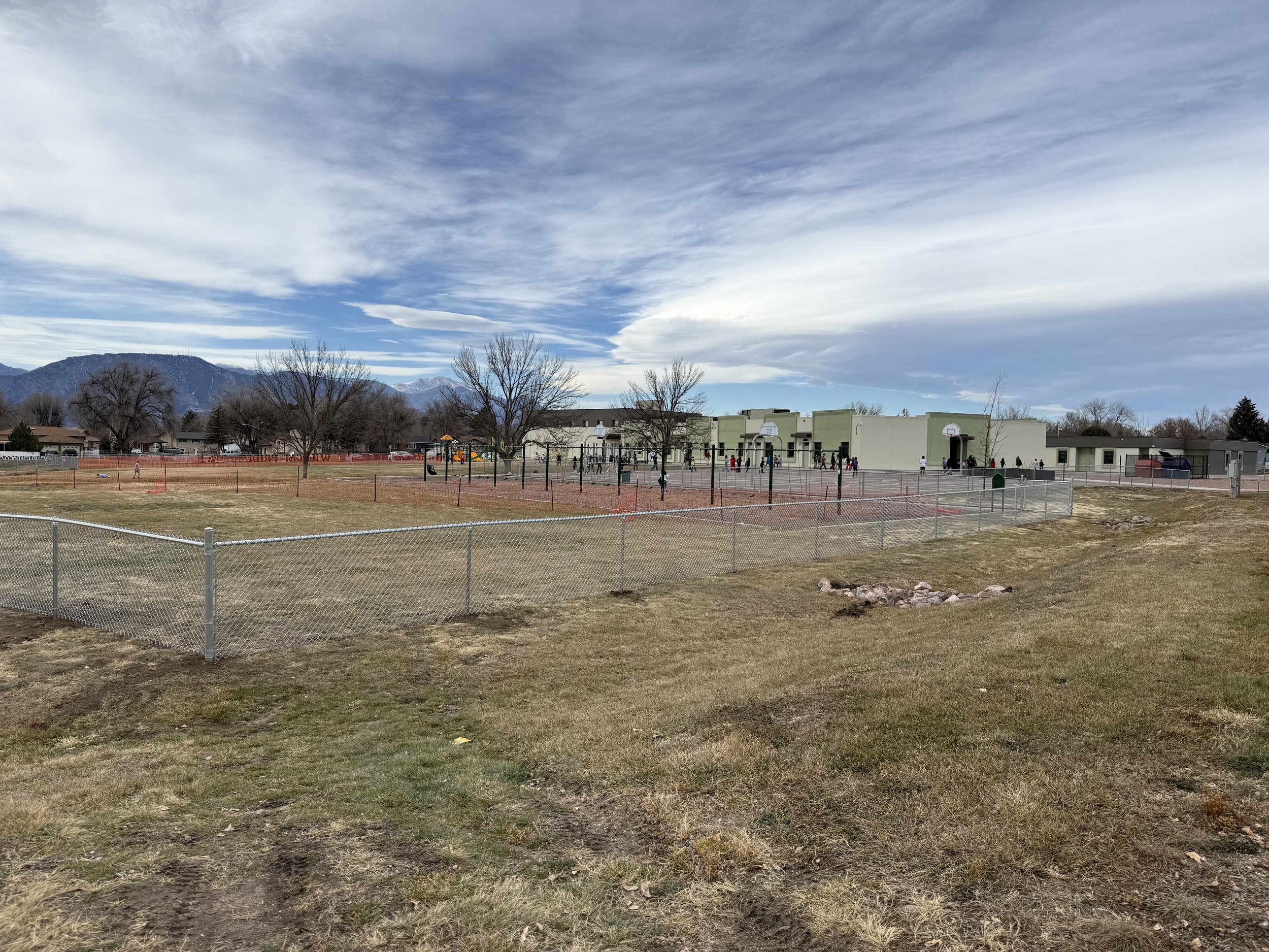 A school playground with a large building, fenced sports courts, leafless trees, and distant mountains under a partly cloudy sky.