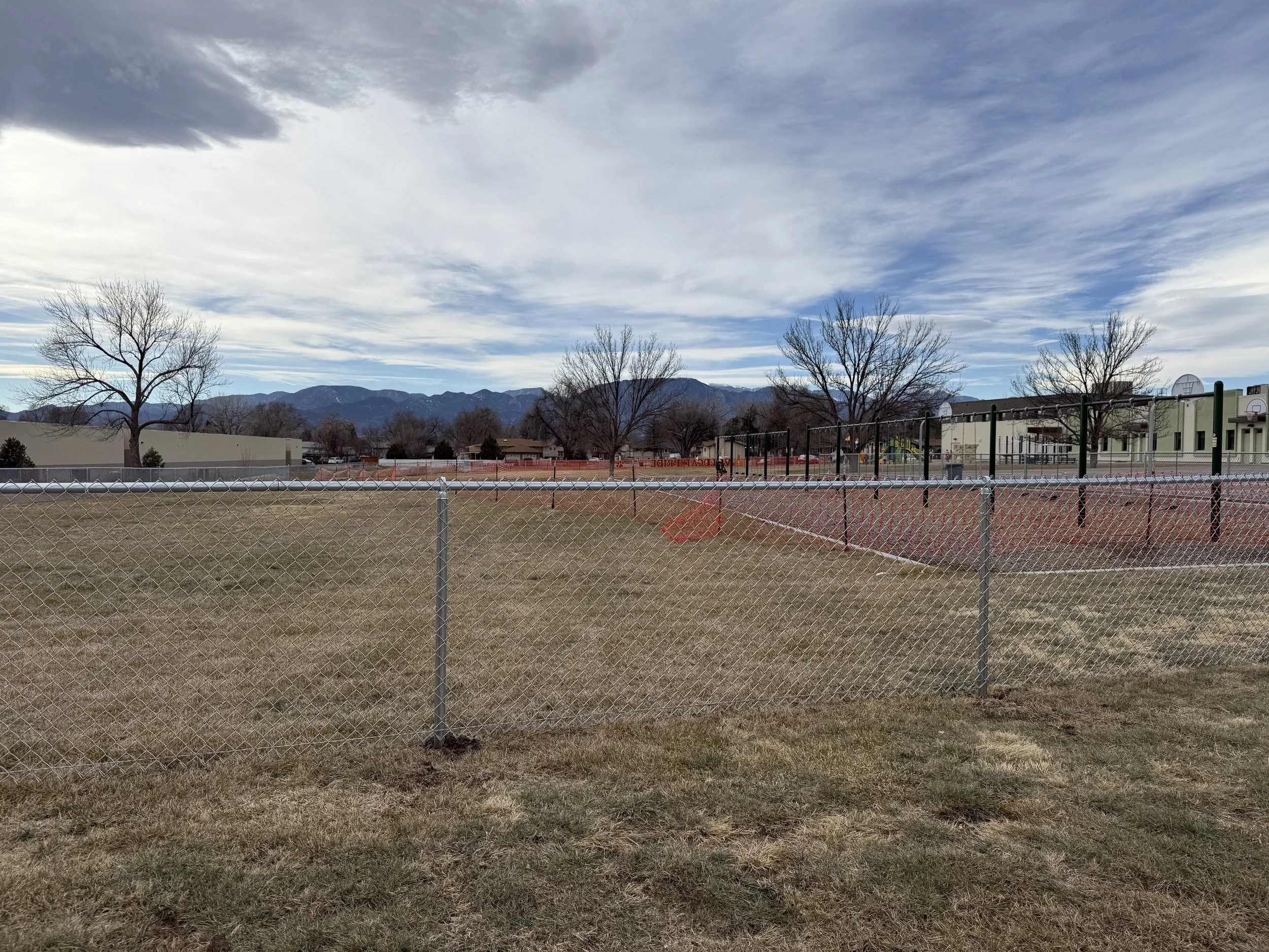 Fenced baseball courts in a park with dry grass, leafless trees, and a mountain range in the background under a partly cloudy sky.