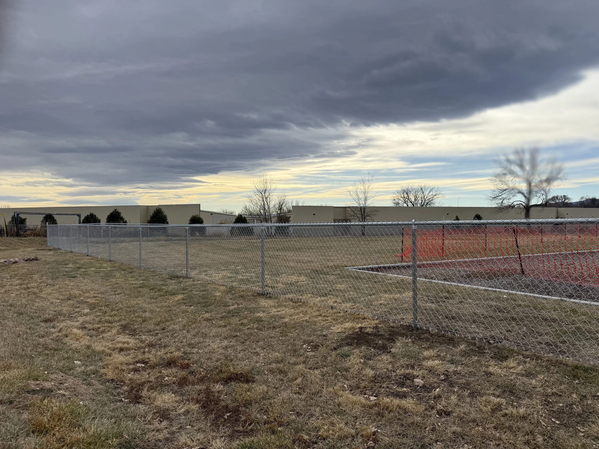 A chain-link fence surrounds an empty grassy area with some leafless trees. Industrial buildings and a cloudy sky with hints of sunlight are in the background.