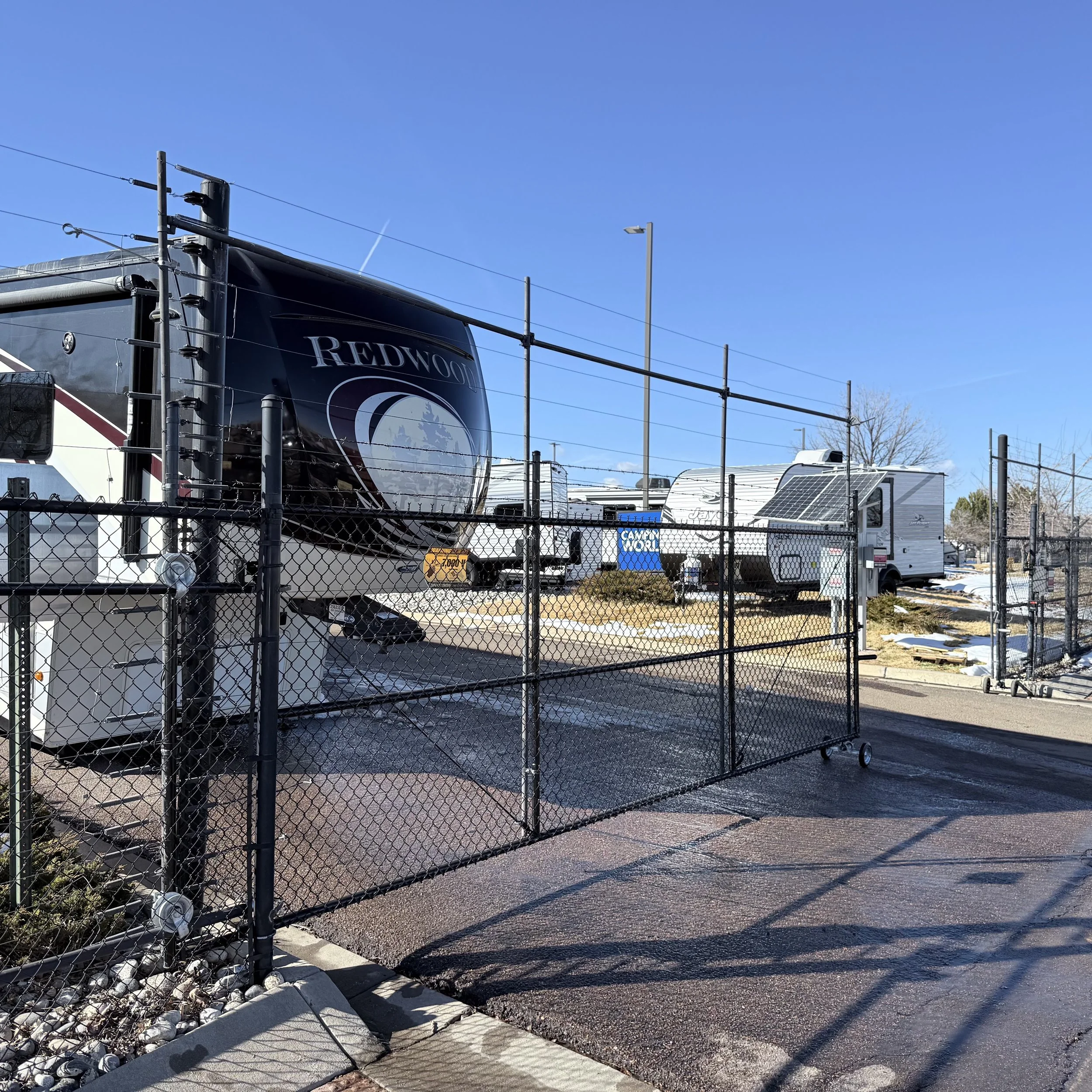 A fenced lot with parked RVs and trailers under a clear blue sky, with snow patches on the ground.
