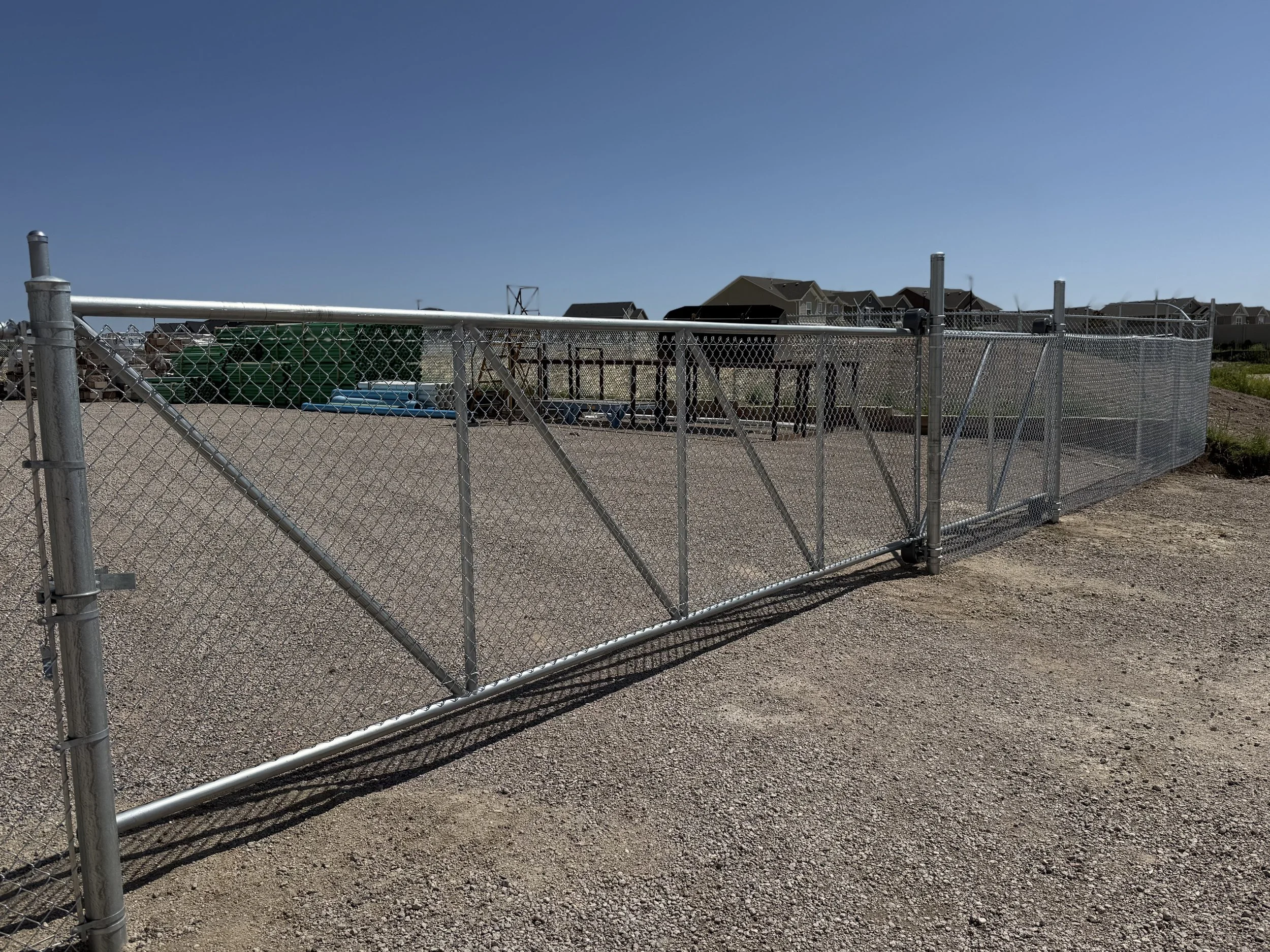 A long metal chain-link fence surrounds a gravel area with a playground or park behind it, under a clear blue sky.