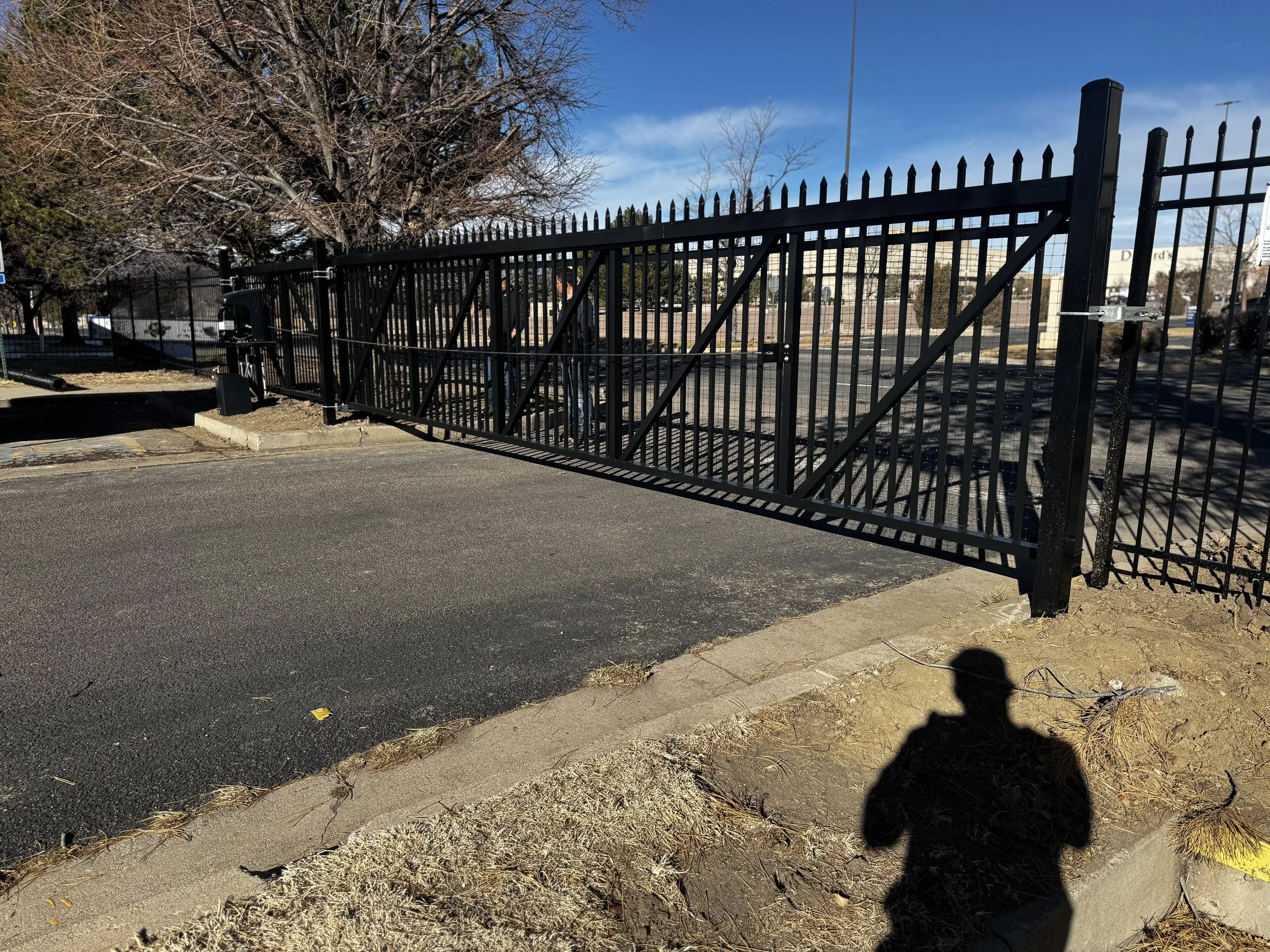 Black metal gate with a lock visible, open in a parking lot, with trees and a building in the background. A person's shadow is cast on the ground.