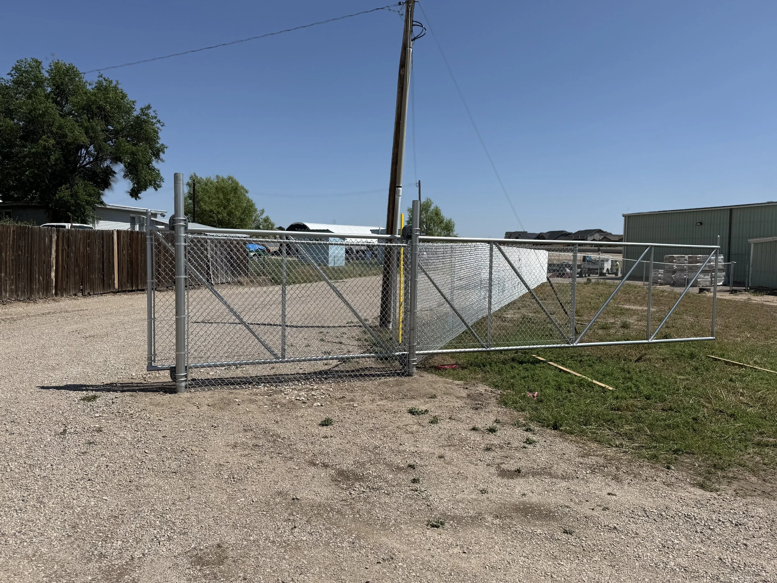 A chain-link fence gate partially knocked over in an outdoor area with gravel and grass. There is a utility pole with wires, a wooden fence, and a building on the right side.