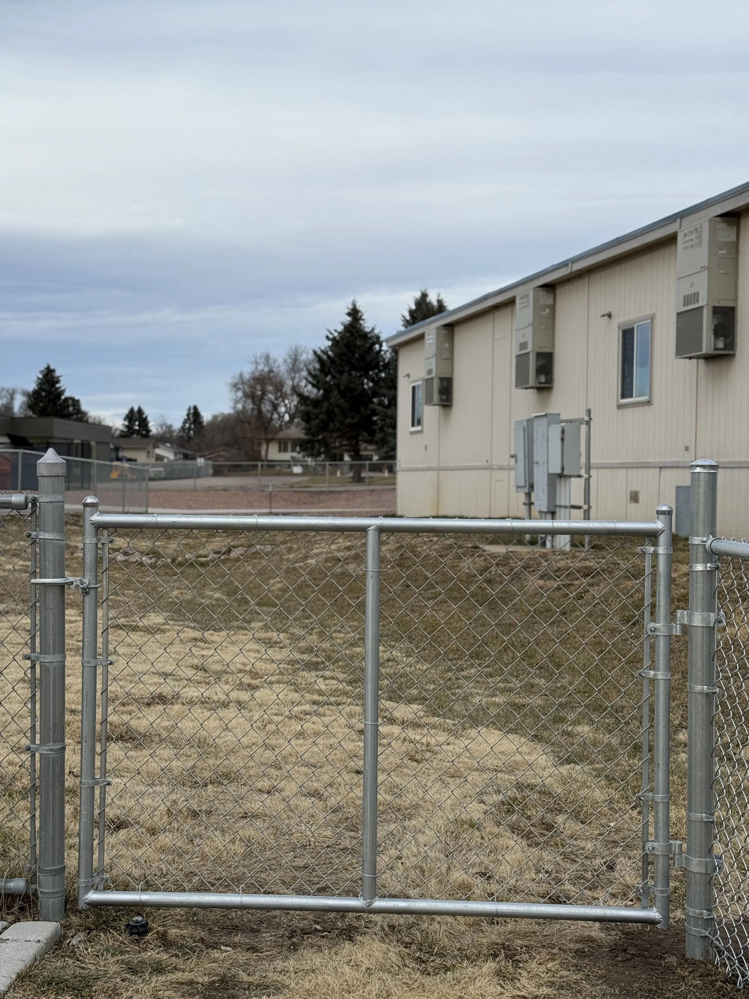 A small metal chain-link gate in front of a beige mobile home with several air conditioning units mounted on the side, and a cloudy sky overhead.