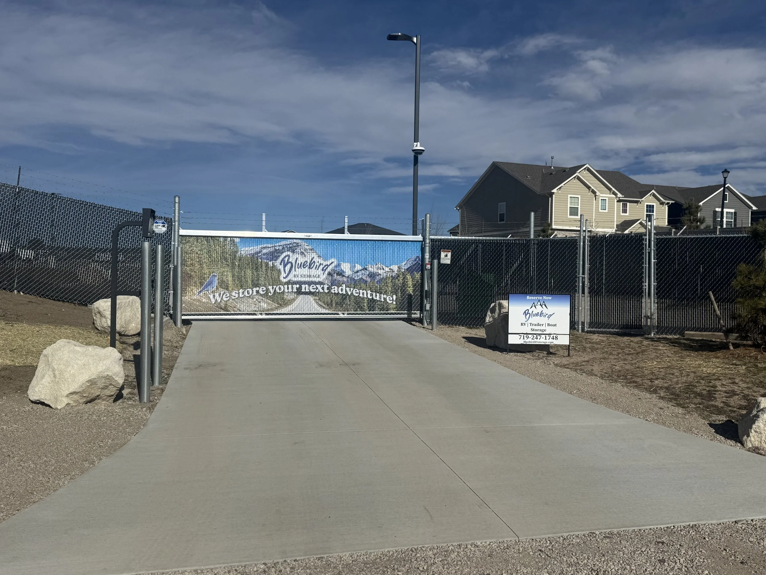 A gate at Bluebird RV Storage with a sign that says 'West store your next adventure!' in front of residential houses under a cloudy blue sky.