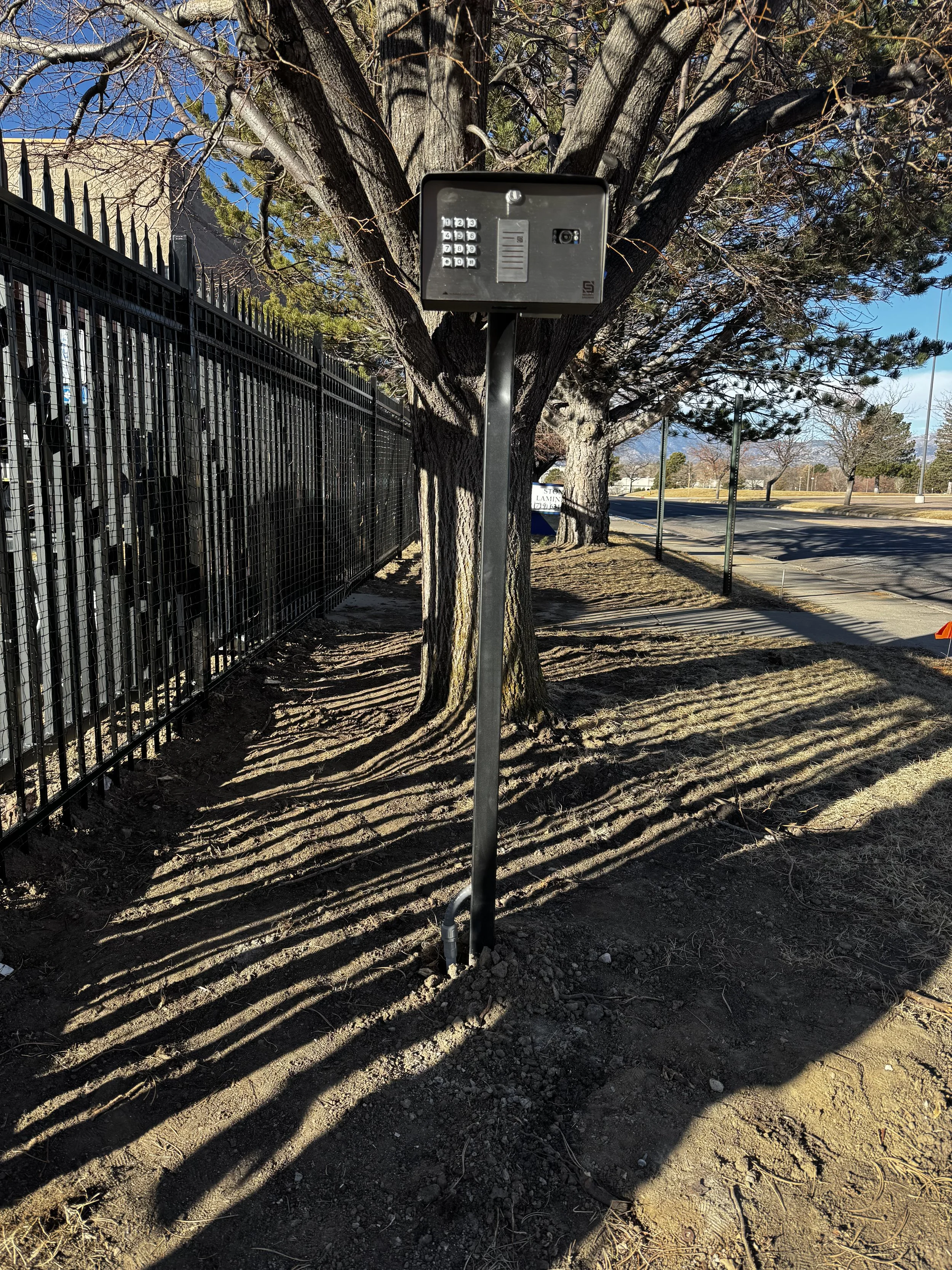 Security keypad and intercom system mounted on a pole next to a tree, casting shadows on the ground, along a sidewalk with a metal fence on the left and a street on the right, on a sunny day.