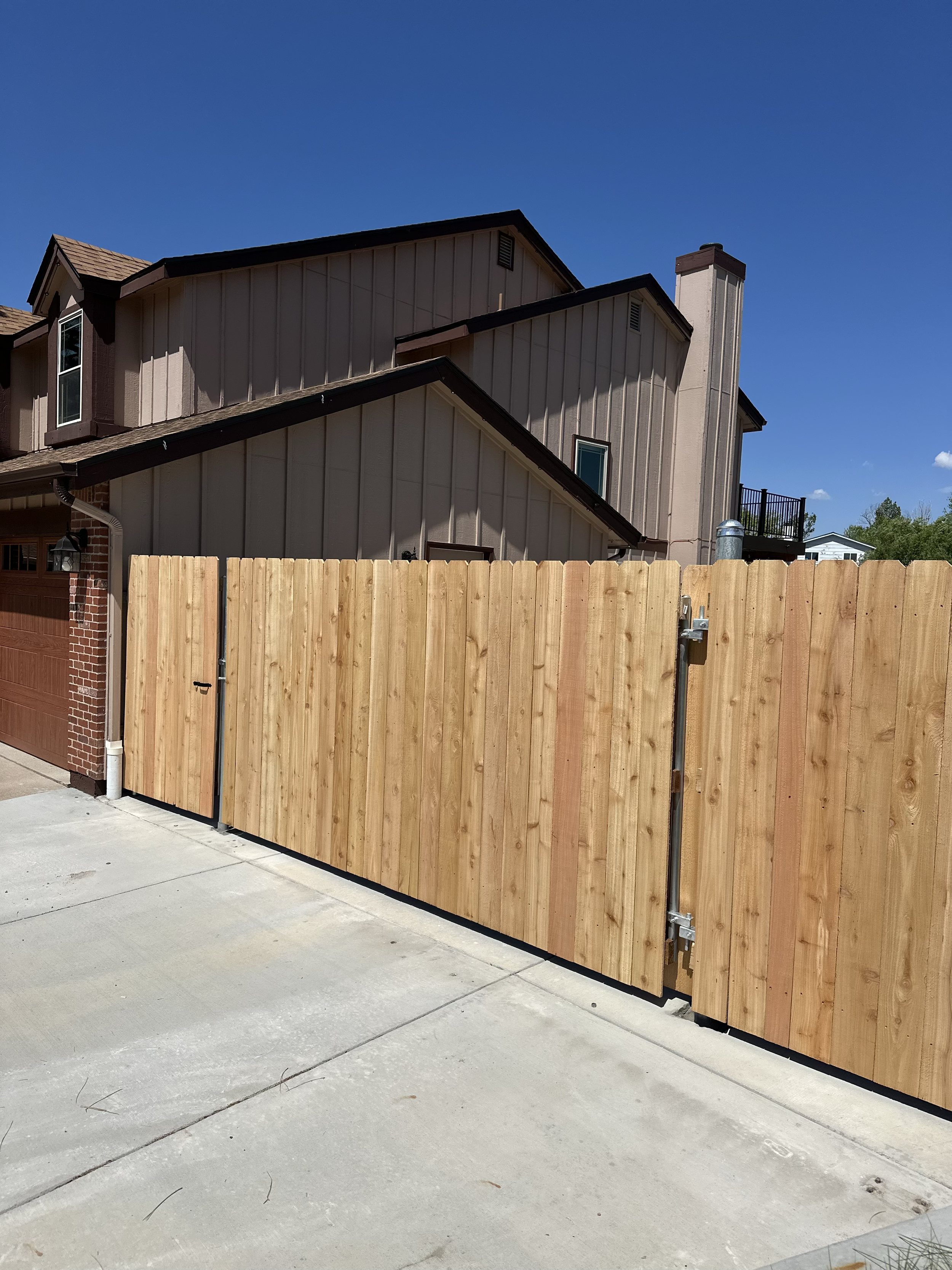 Wooden privacy gate and fence in front of a house with beige siding and a brown garage door, under a clear blue sky.