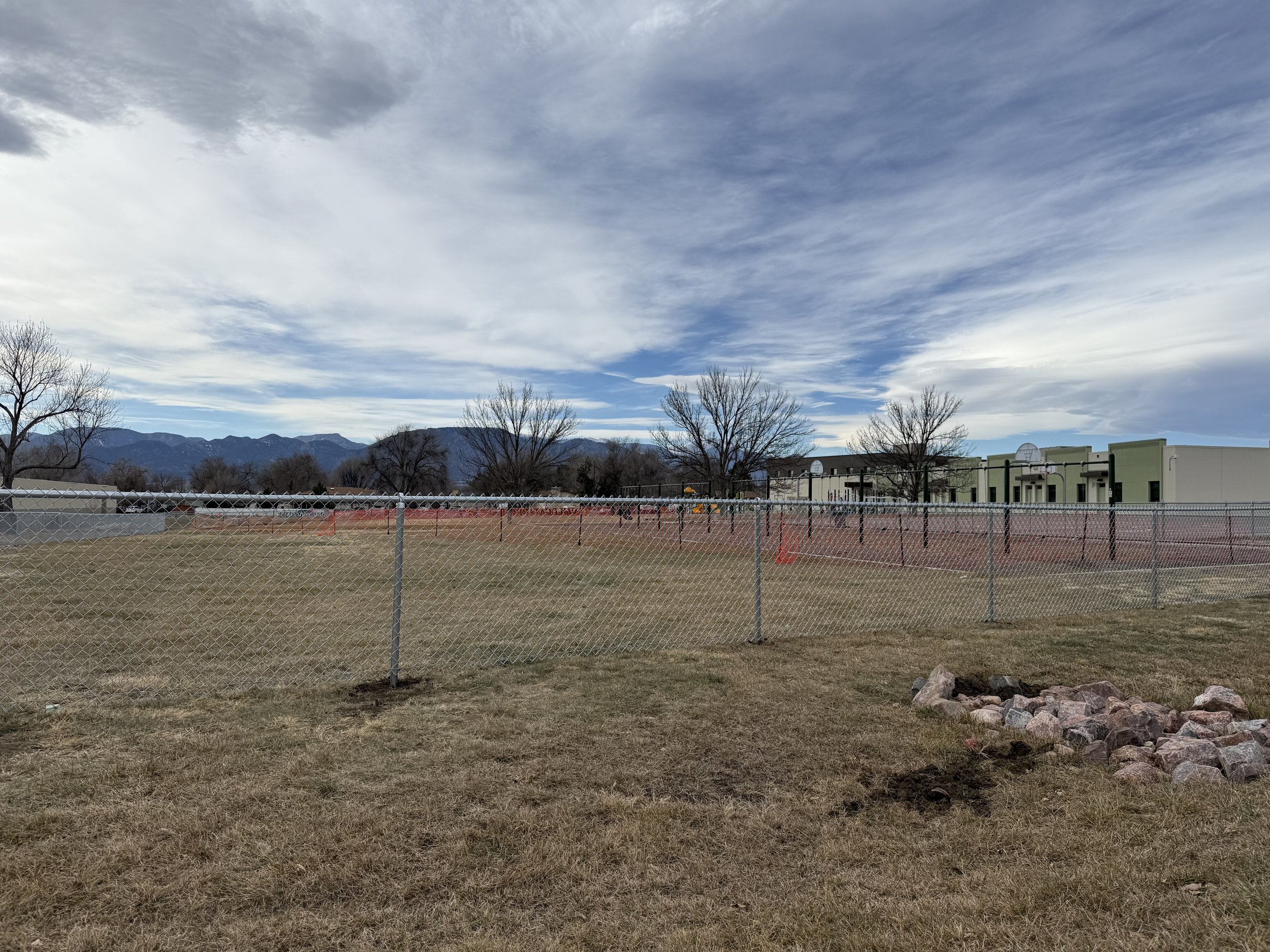 Open grassy field with a chain-link fence, some small trees, a row of modern buildings, and mountains in the background under a partly cloudy sky.