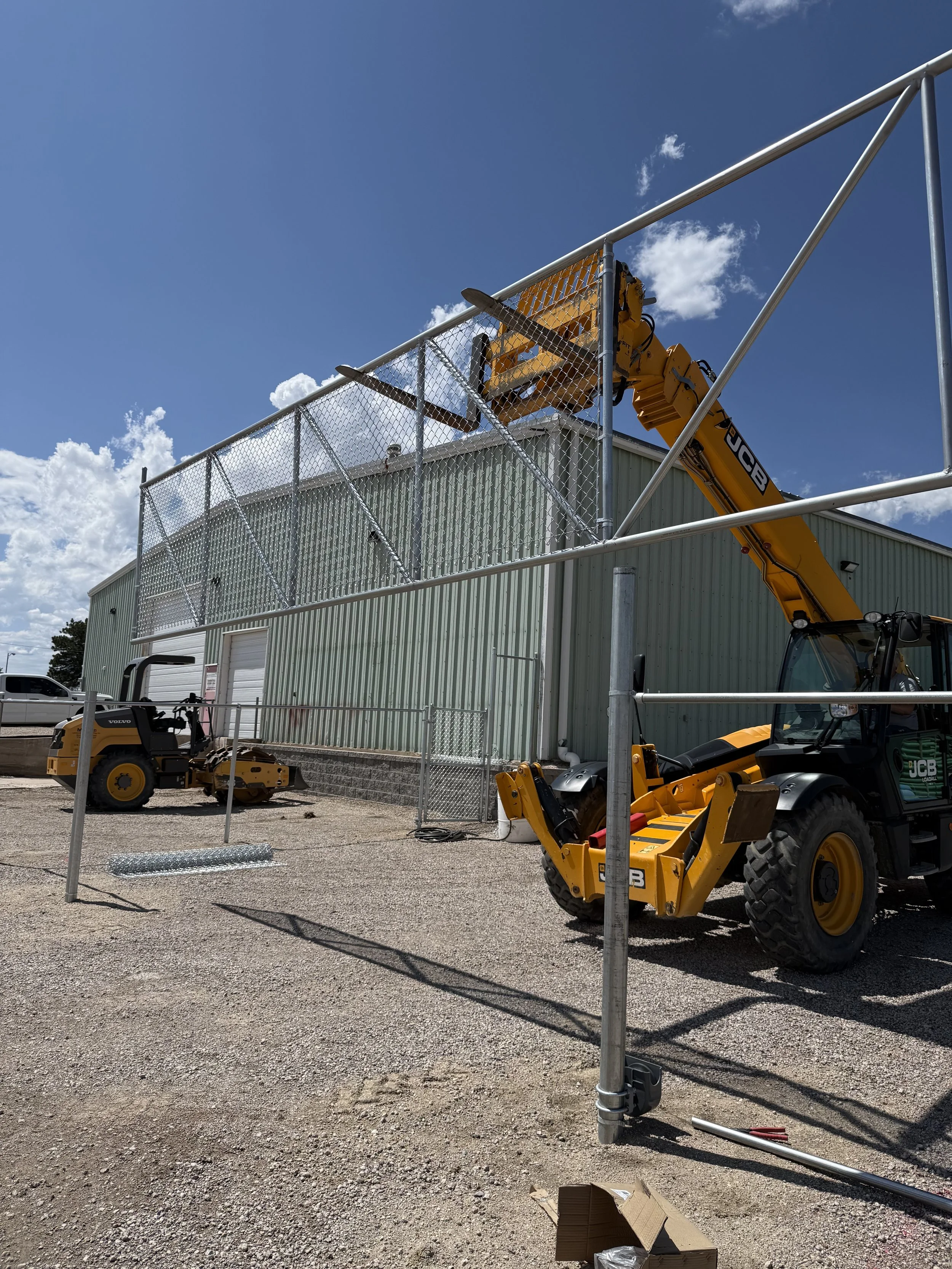 Construction site with a green building, metal scaffolding, and two yellow cranes lifting materials. The area is gravelly with some tools and equipment visible.