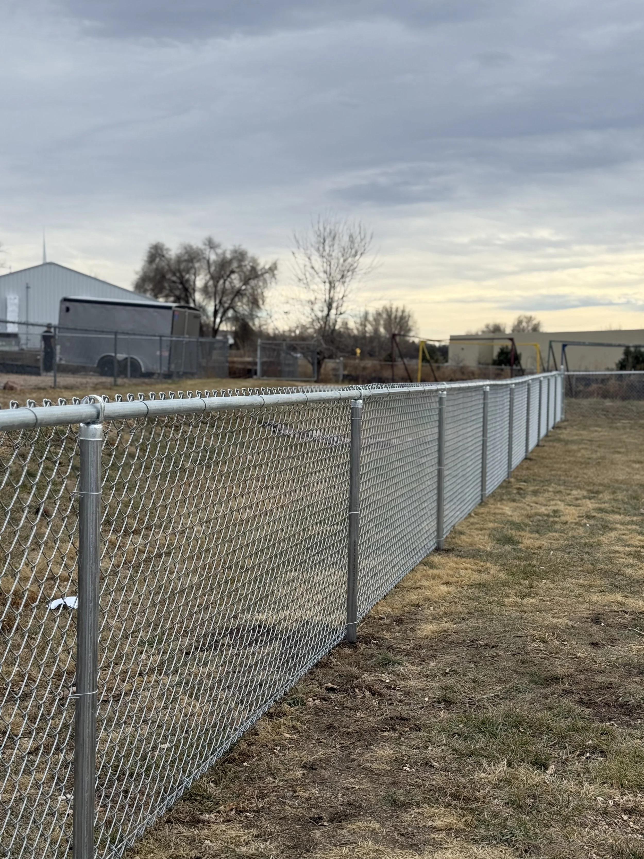 A chain-link fence running alongside a grassy field with a playground in the background, under a cloudy sky.
