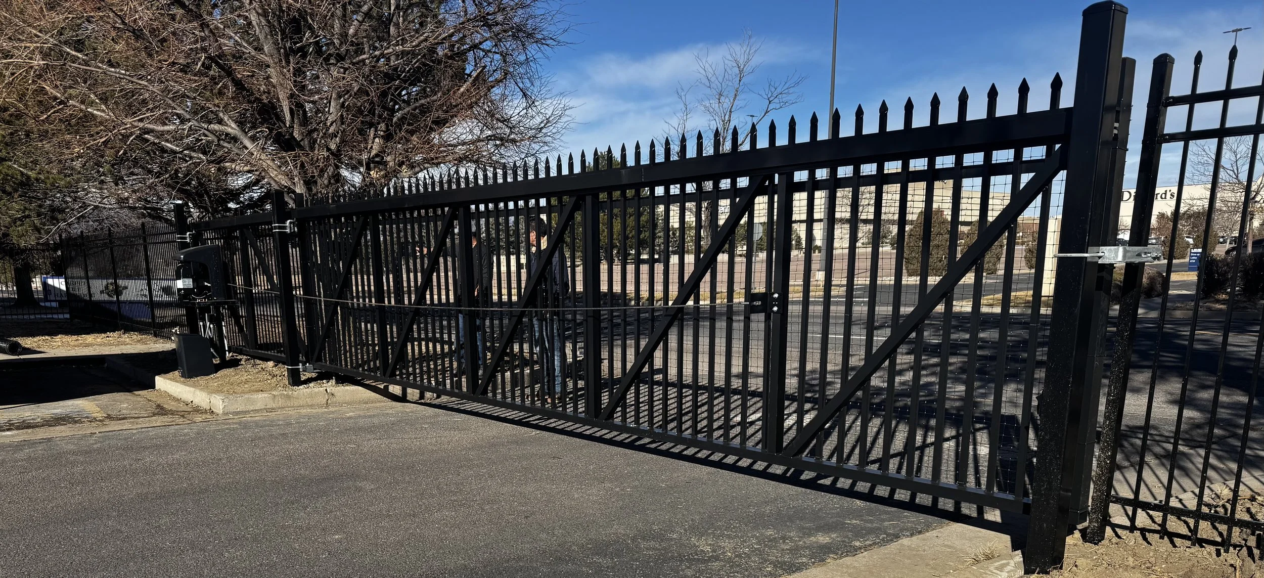 Black metal driveway gate with diagonal bars, partially open, showing a parking lot and trees in the background.