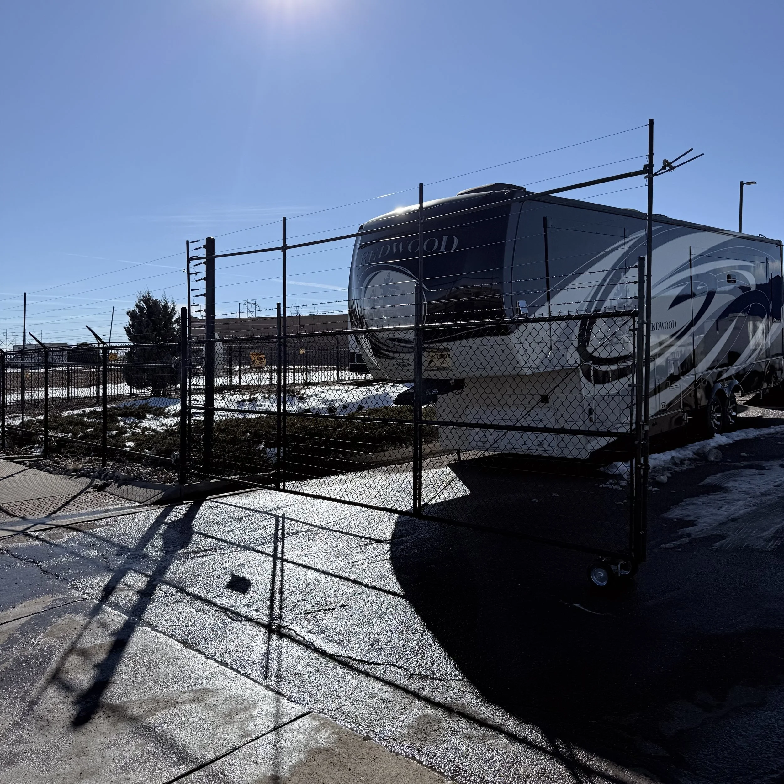 A large RV parked behind a black chain-link fence on a sunny day with patches of snow on the ground.