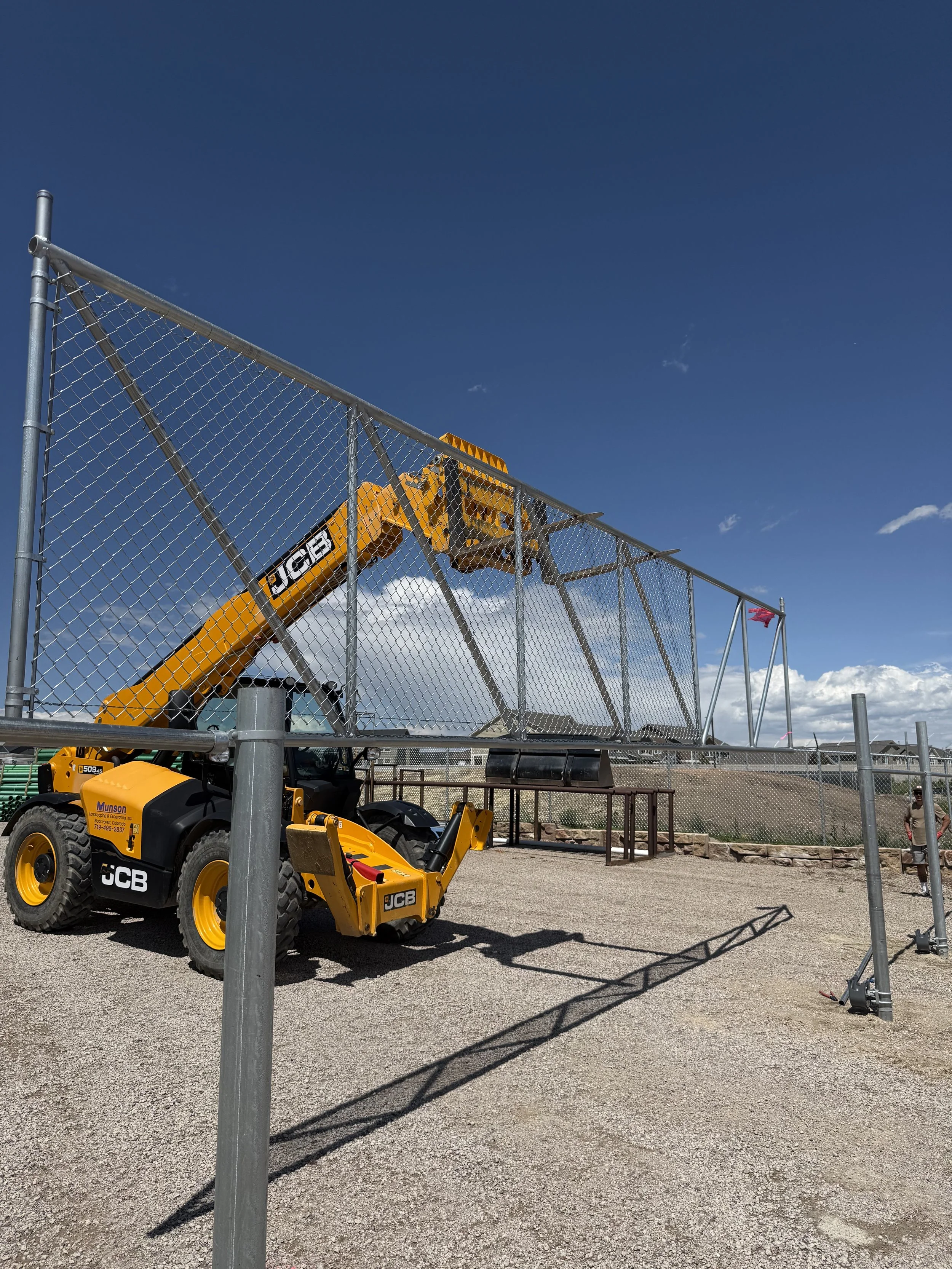 A construction crane is installing a chain-link fence around a construction site on a sunny day with blue sky and scattered clouds.