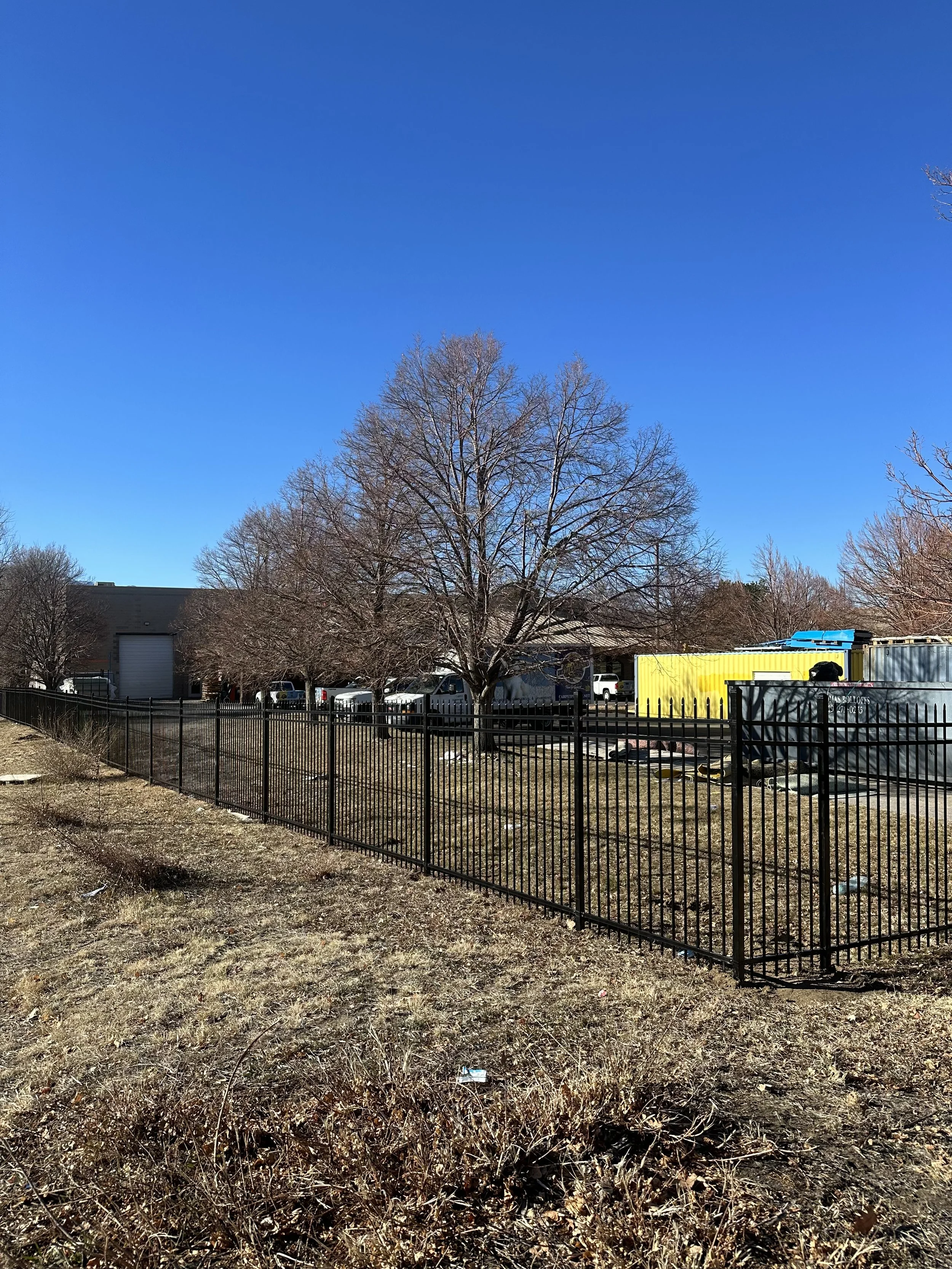 A black metal fence runs along a patch of dry grass, with leafless trees and parked trucks behind it under a clear blue sky.