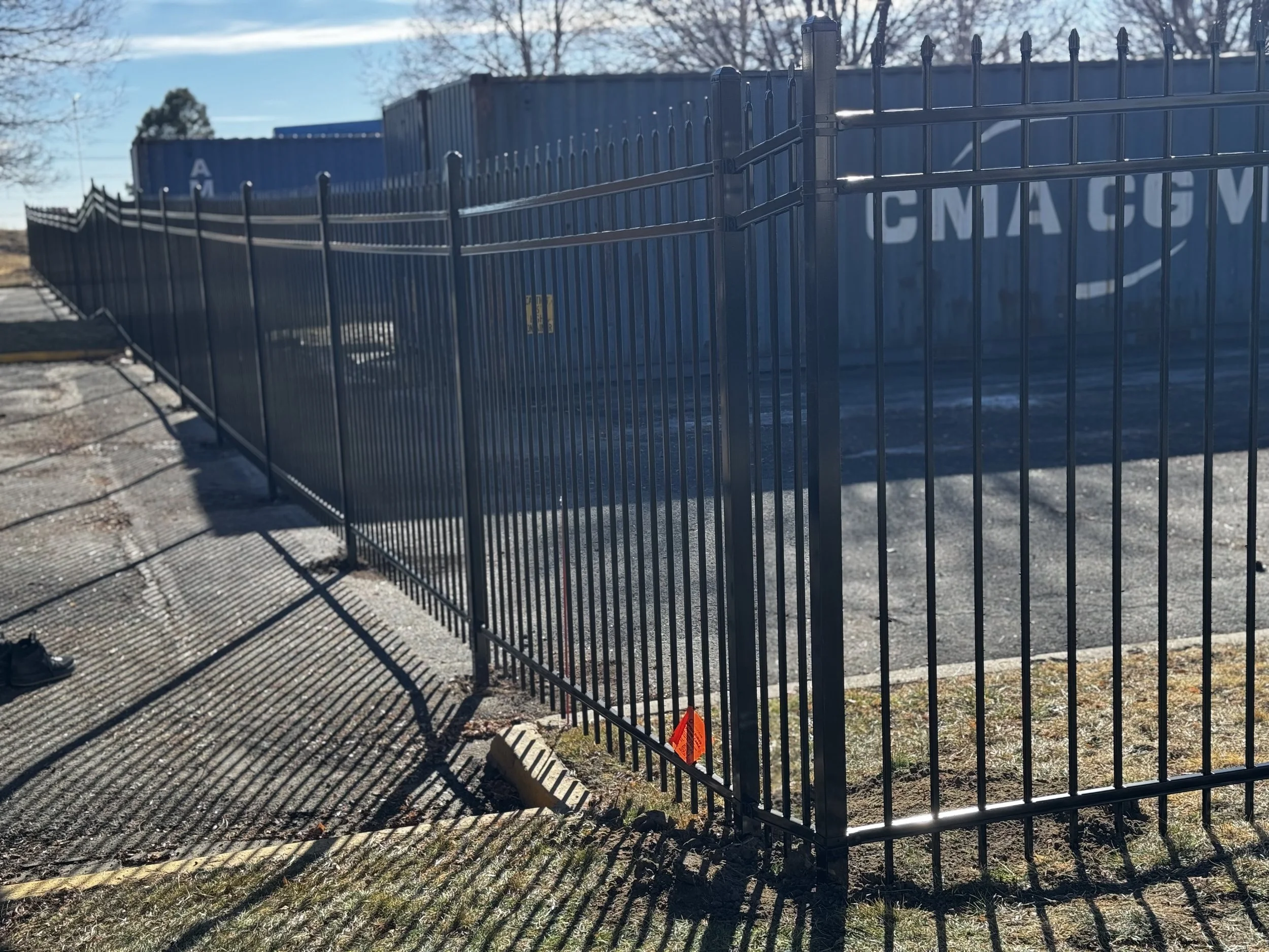 Black metal fence surrounds a storage area with blue shipping containers, casting shadows on the ground. The sky is clear and the scene is well-lit.
