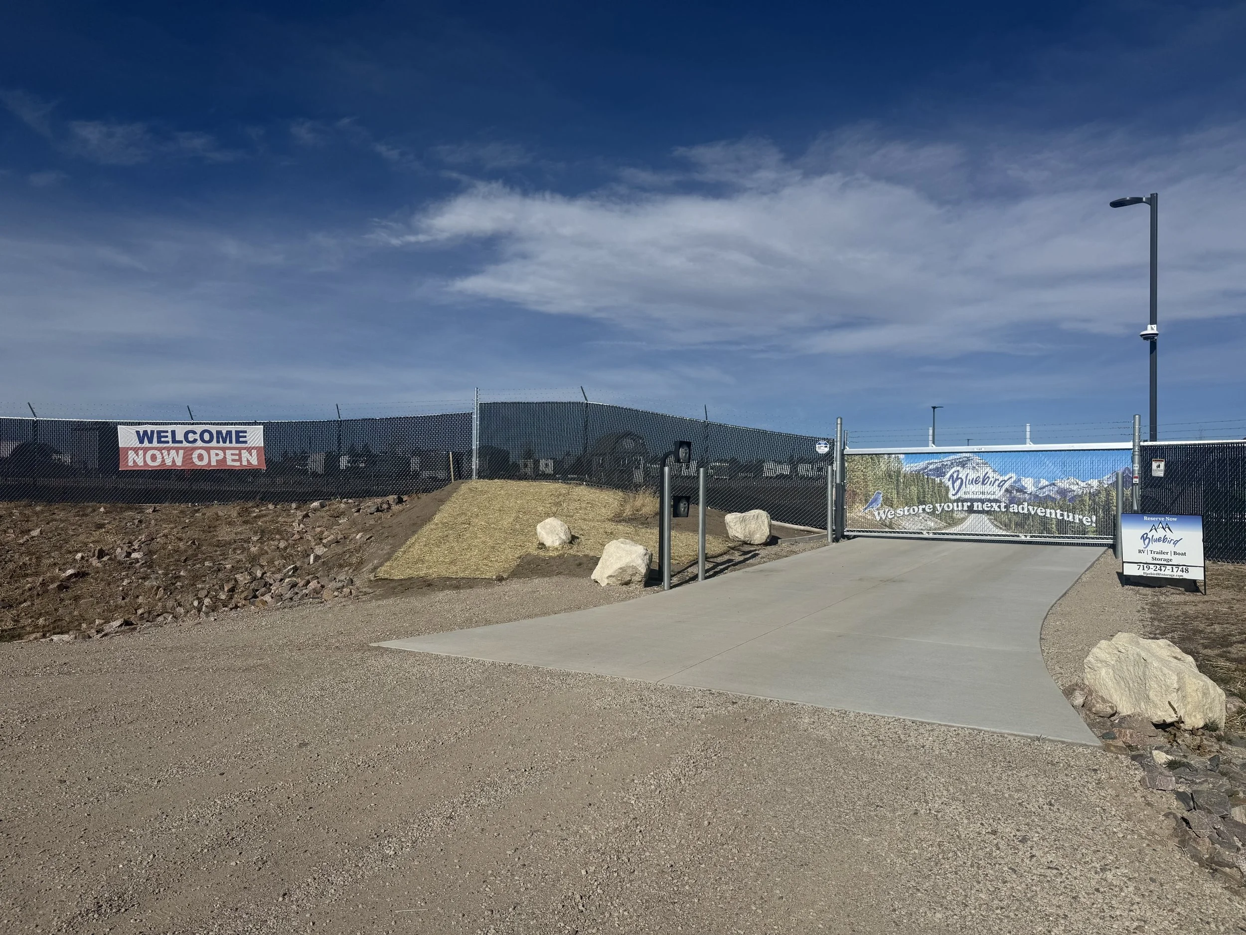 Entrance to a Bluebird RV Resort with a concrete road, large rocks, and signs that read "Welcome Now Open," "Bluebird," and "We store your next adventure!" under a blue sky with scattered clouds.