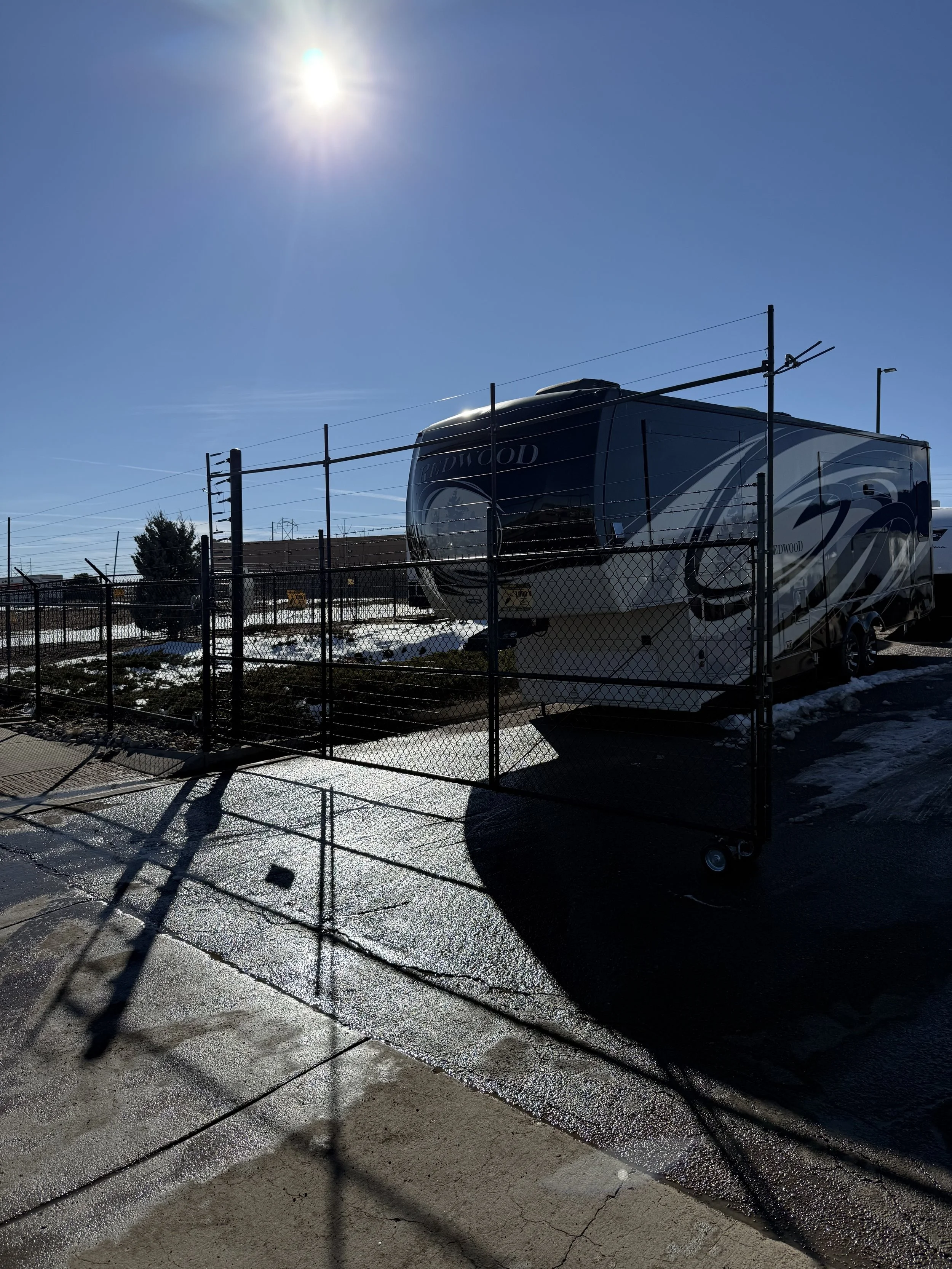 A parked RV behind a black chain-link fence on a sunny winter day, with snow on the ground and shadows cast by the fence.