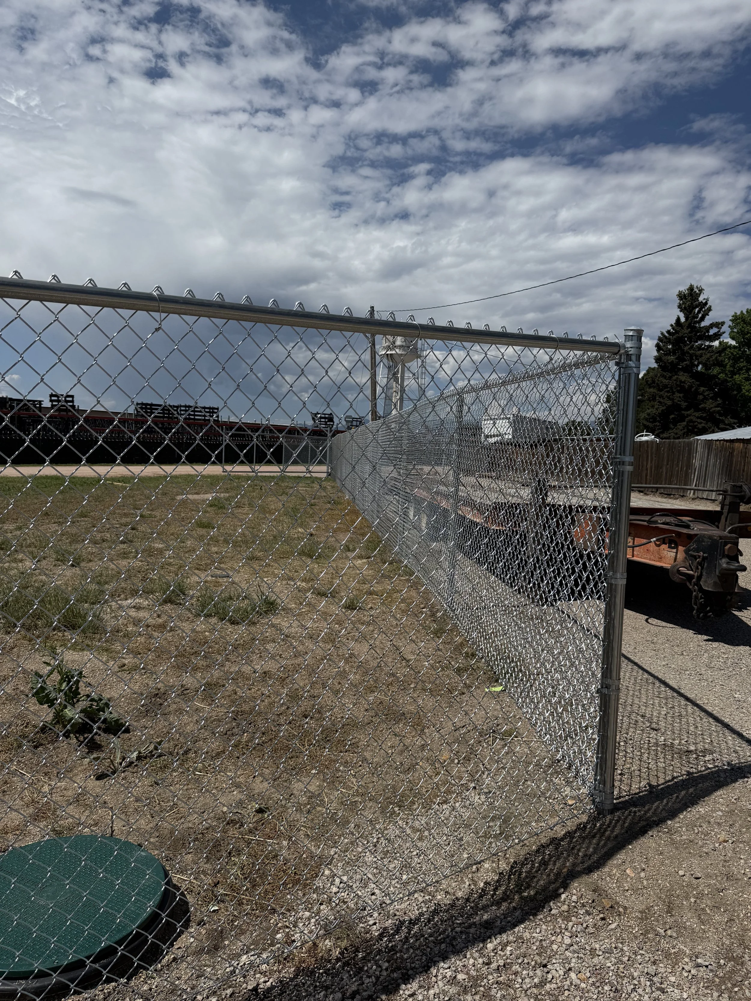 Metal chain-link fence with an open gate, on a grassy area with a cloudy sky overhead and some trees and equipment in the background.