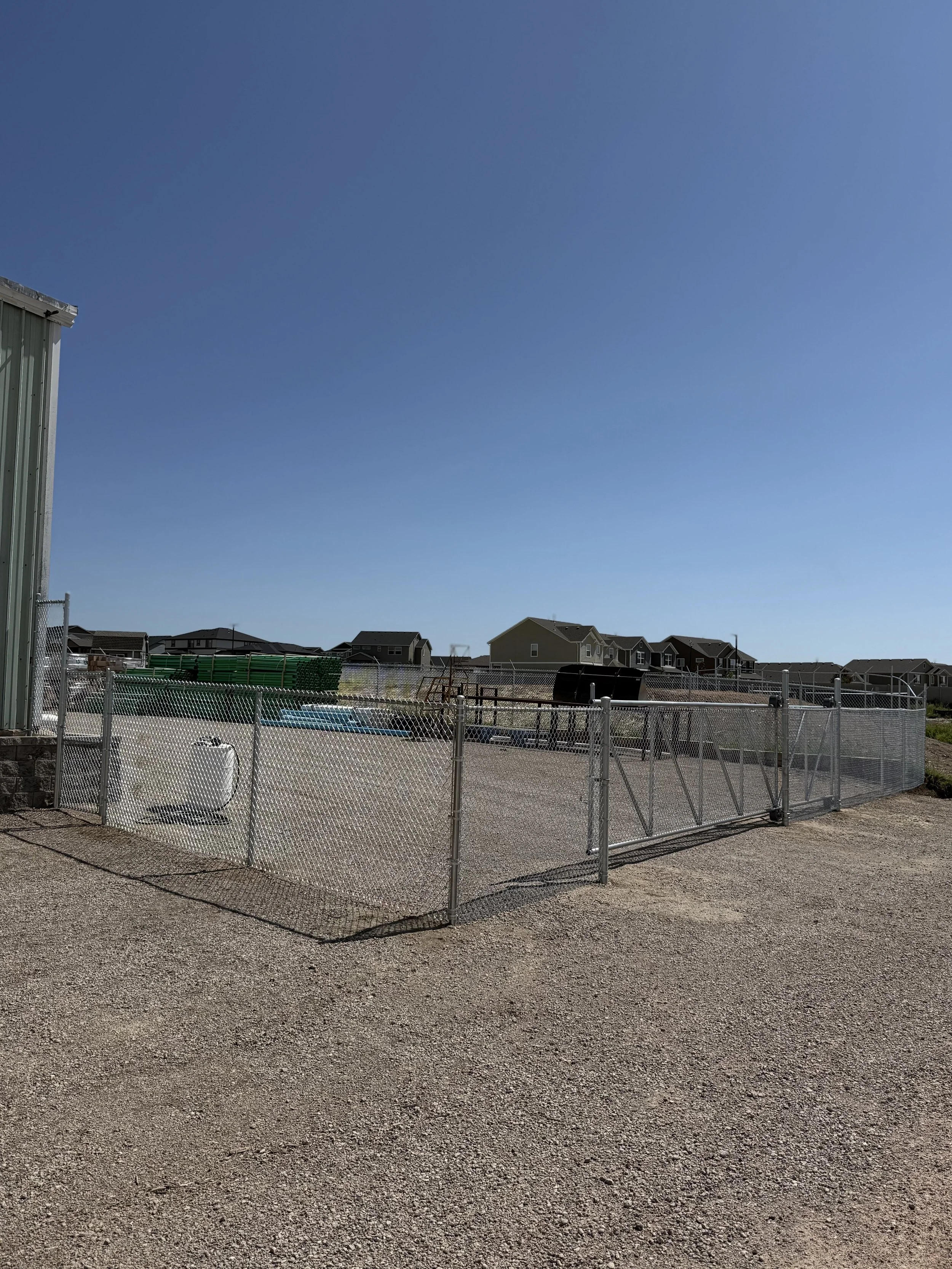 A fenced outdoor area with gravel ground, a metal shed on the left, and residential houses in the distance under a clear blue sky.