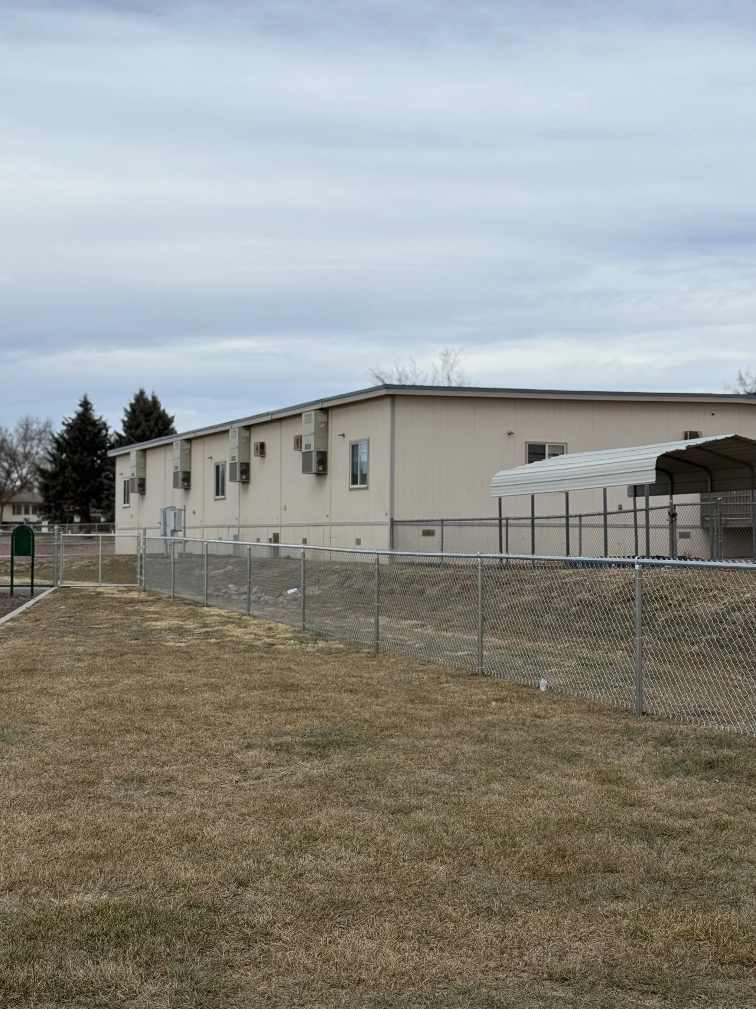 A beige apartment building with several air conditioning units on the exterior and a chain-link fence in the foreground, surrounded by a grassy yard under a cloudy sky.