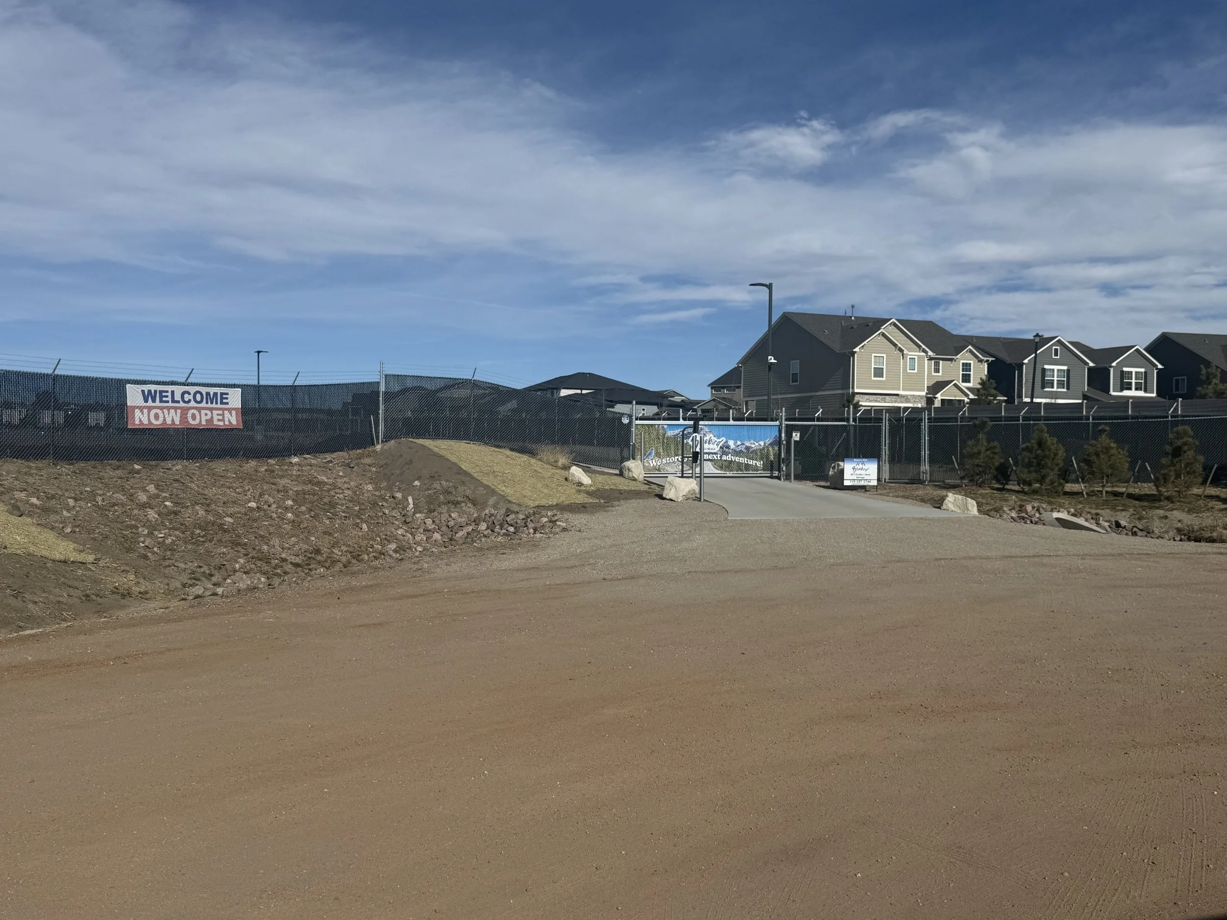 Entrance to a new residential neighborhood or housing development with a fence, a 'Welcome Now Open' sign, a paved pathway, and large rocks along the dirt area.