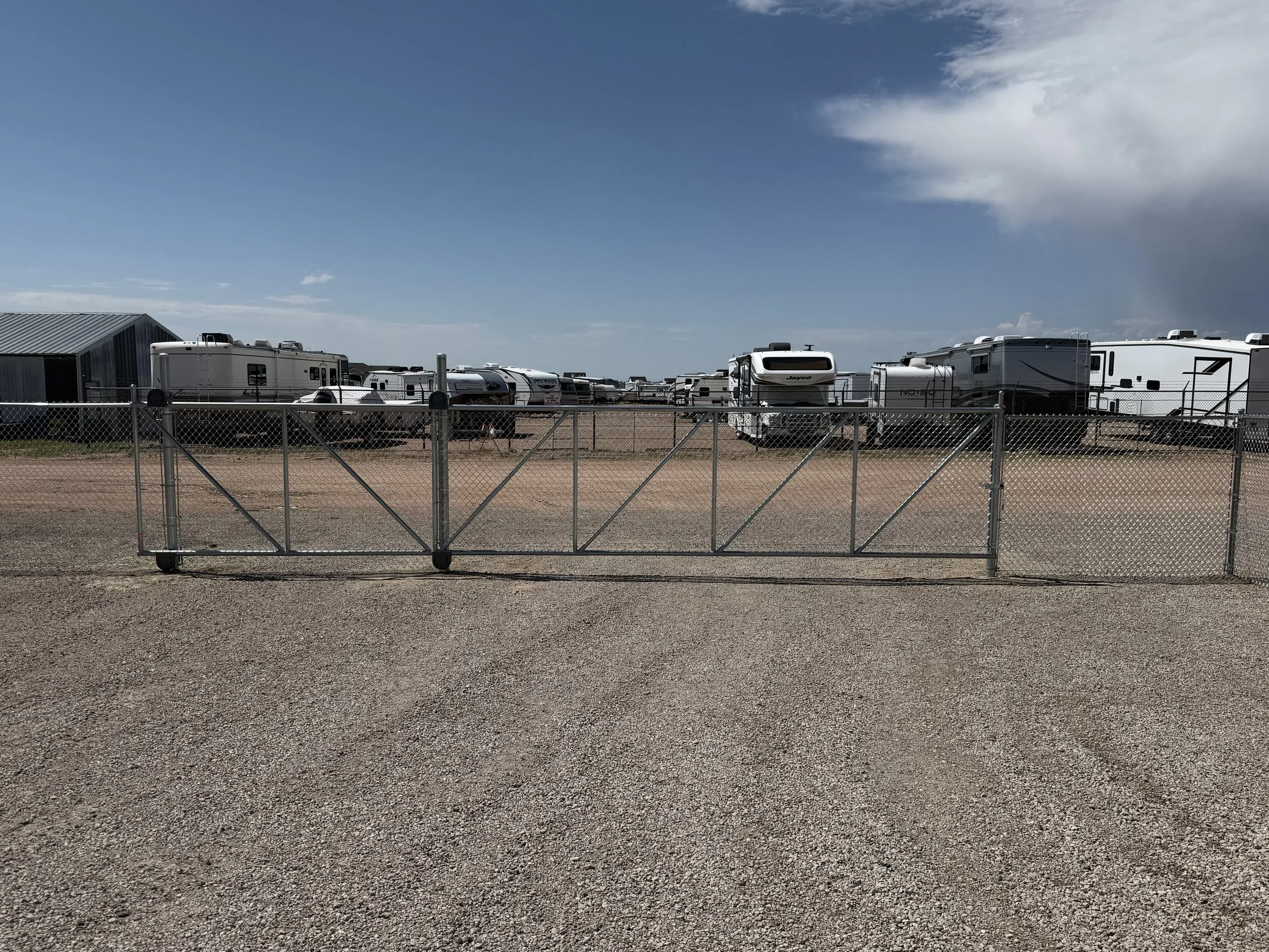 A fenced area with a metal gate in front, containing numerous recreational vehicles (RVs) parked on a dirt lot. The sky is partly cloudy.