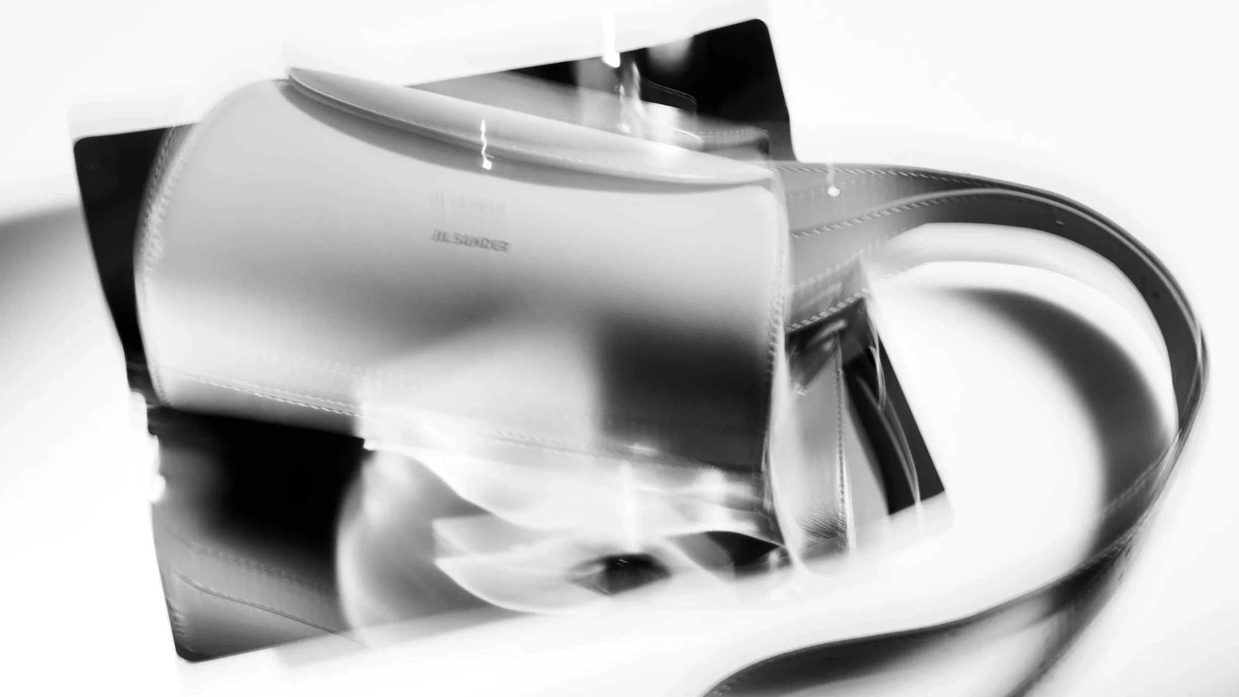 A reflective black-and-white photograph of a white leather handbag with a curved handle, placed on a mirror surface, creating a mirrored reflection of the bag and its handle.