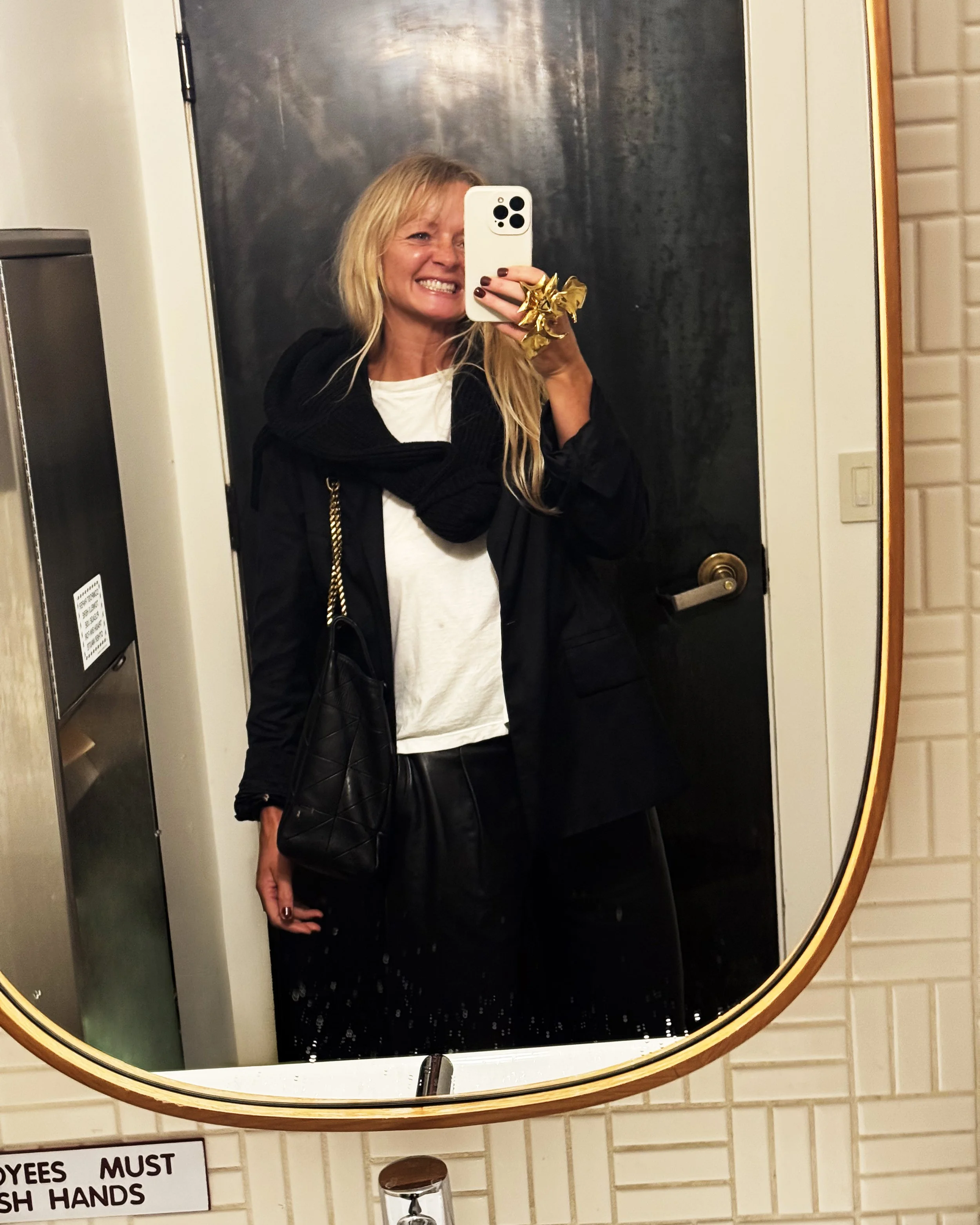 A woman taking a selfie in front of a mirror in a bathroom, smiling and showing her teeth, wearing a black jacket over a white shirt, with a large gold bow ring on her finger, holding a black quilted purse with a gold chain strap.
