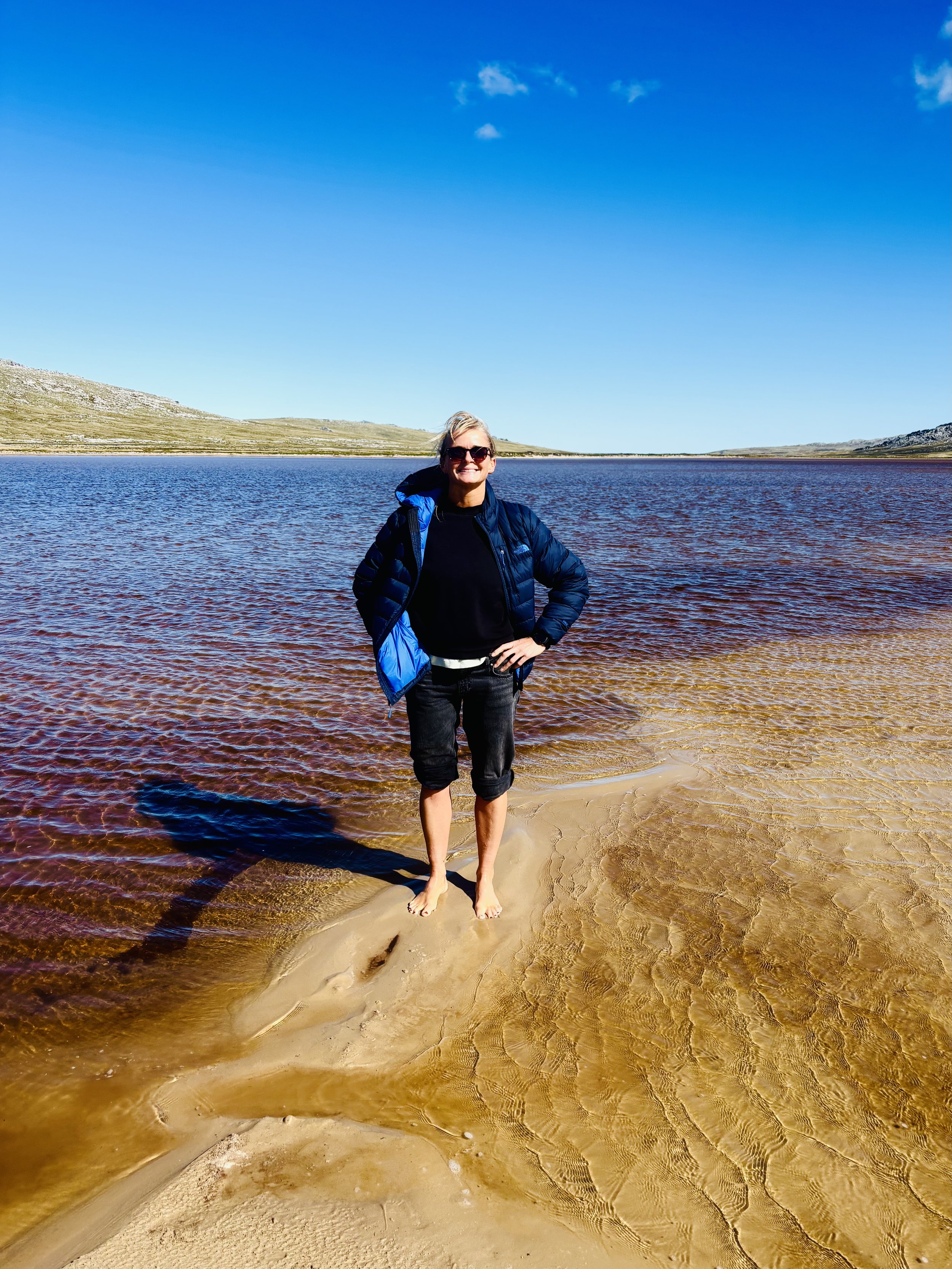 A woman standing barefoot on sandy shore by a large lake with red-tinted water, wearing a blue jacket and black pants, under a clear blue sky with rolling hills in the background.