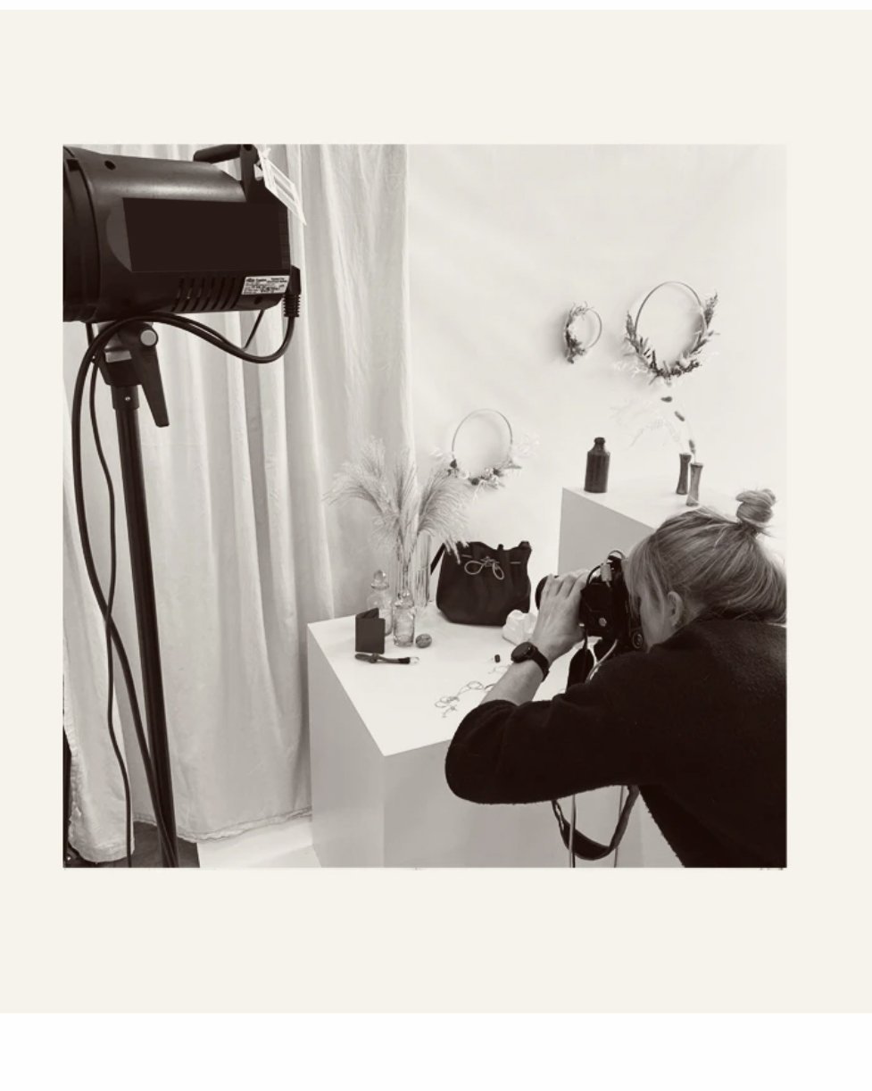 A woman taking photographs in a minimalistic, decorated studio or room with white walls, featuring decorative wreaths, vases, and a black bag on a white table.