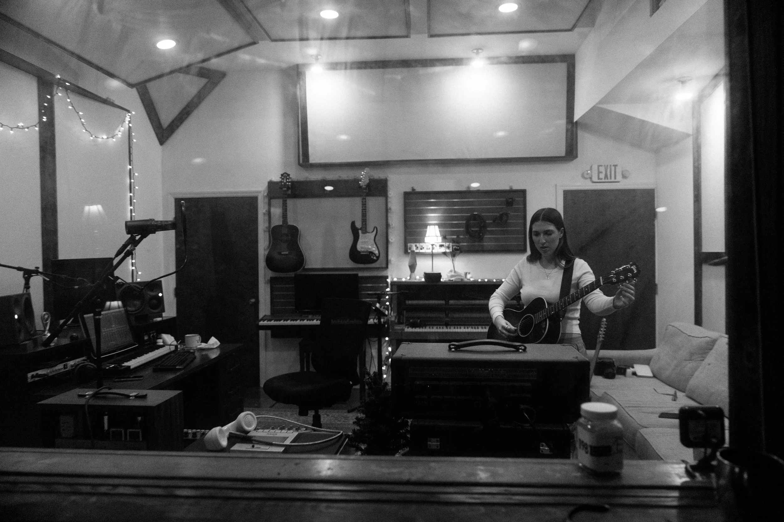 Black and white photo of Julia DeTomaso seen through control room glass, standing with acoustic guitar in a studio surrounded by instruments and warm lighting.