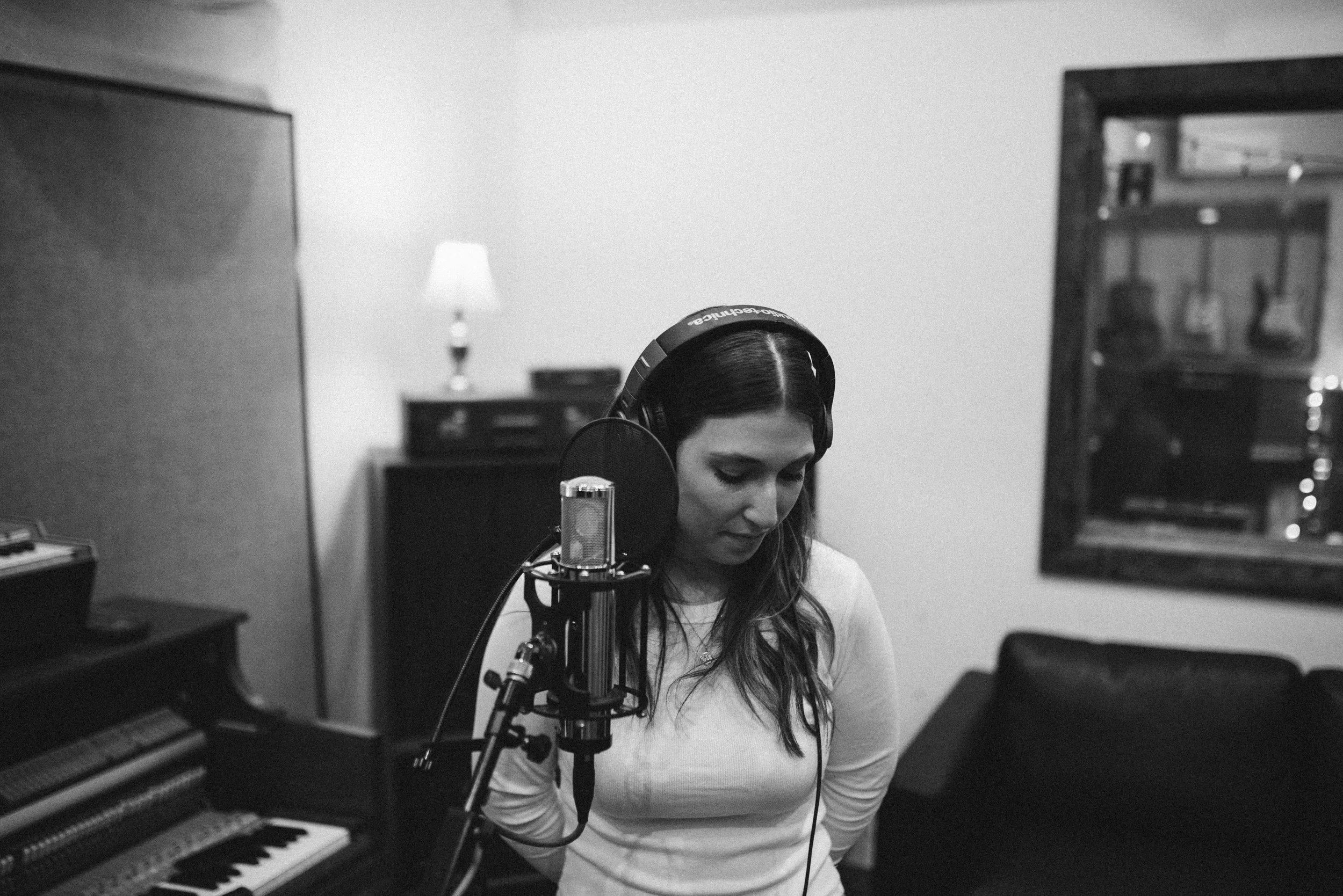 Julia DeTomaso in studio recording vocals — black and white photo with piano and mic setup in background.