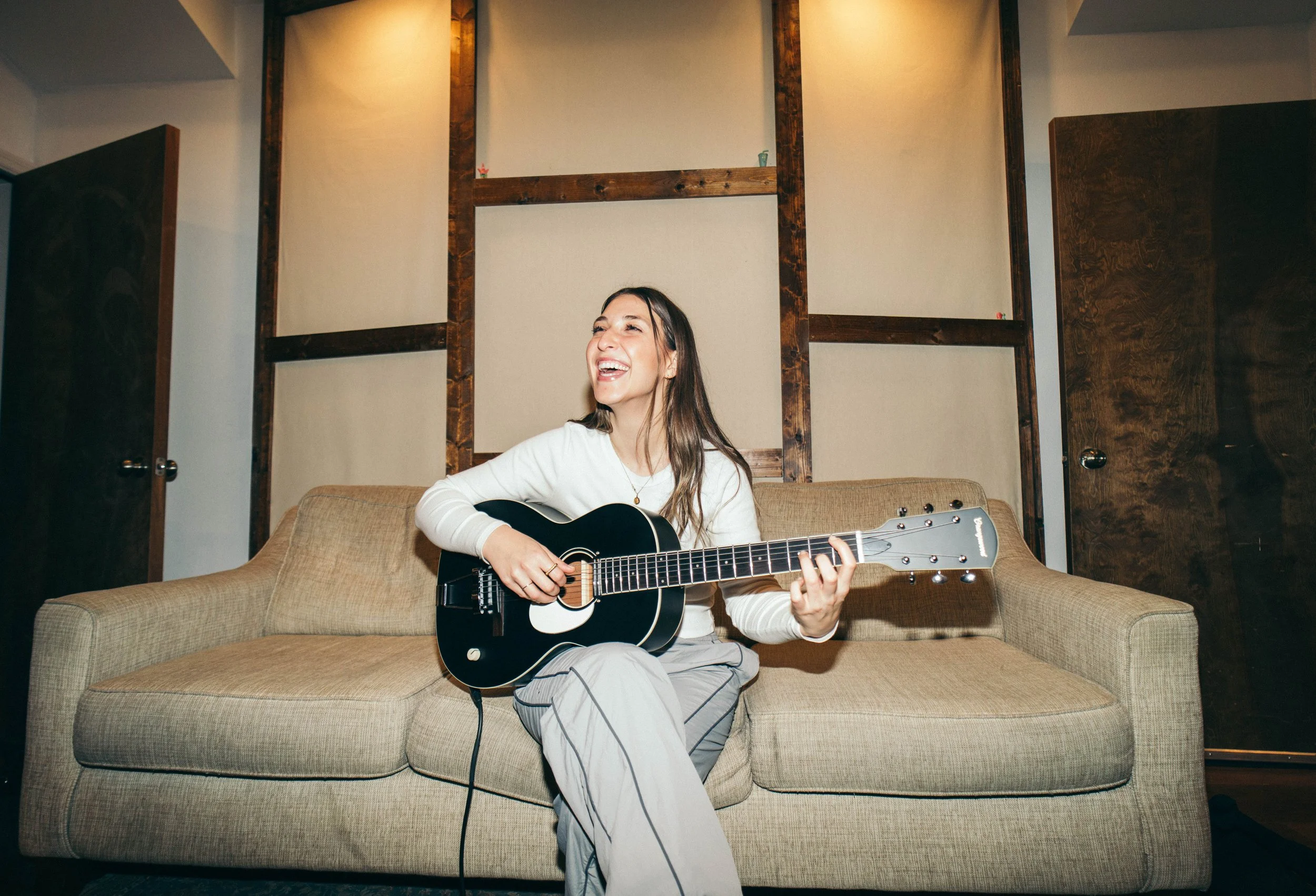 Julia DeTomaso smiling on beige couch with acoustic guitar — warm indoor studio portrait with wood-paneled walls.