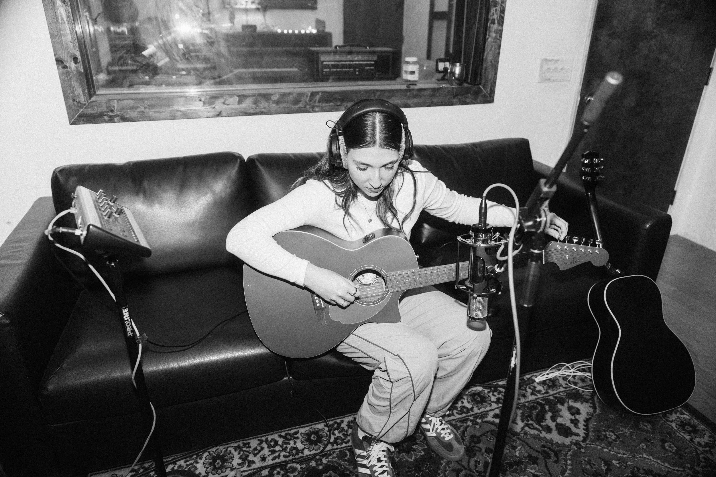 Black and white photo of Julia DeTomaso adjusting studio headphones while recording acoustic guitar on a couch — surrounded by microphones, reflection glass, and a patterned rug.