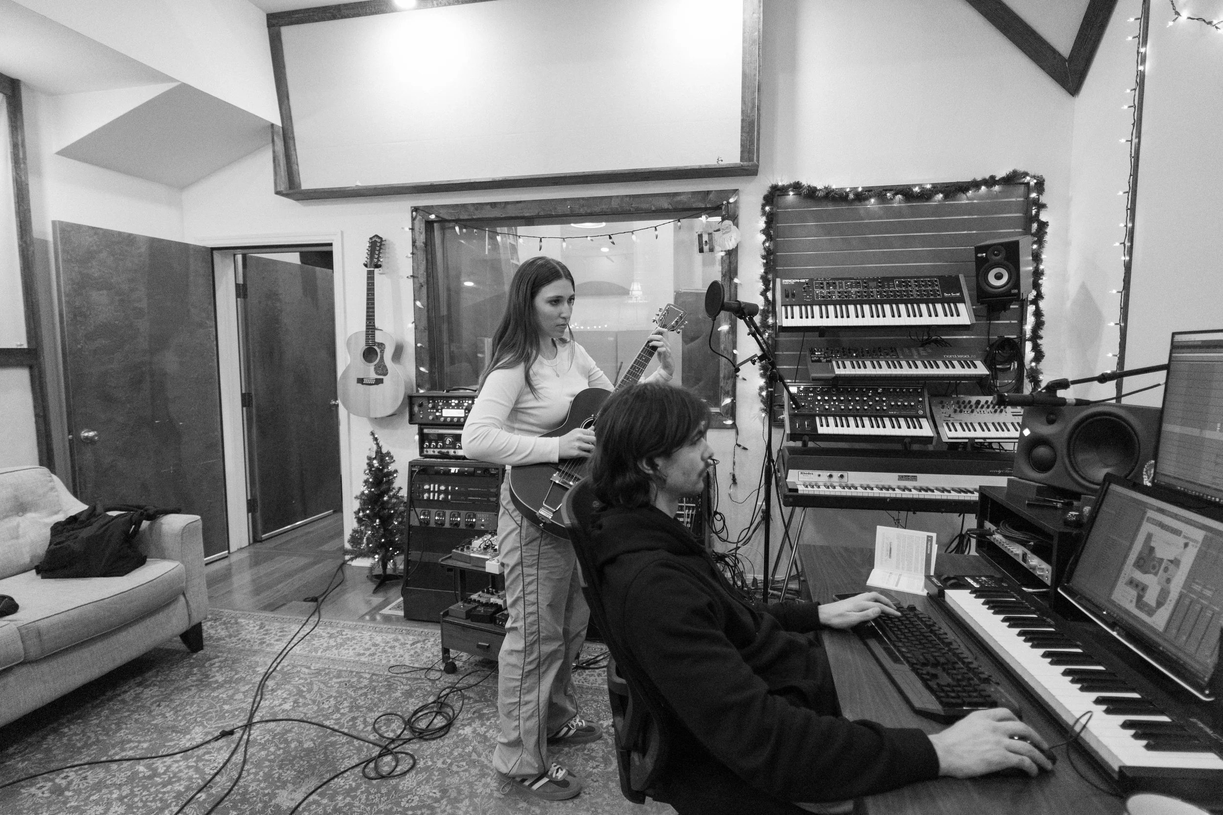 Julia DeTomaso in studio playing electric guitar while producer works at computer — black and white photo with synths, keyboard, and guitars in the background.