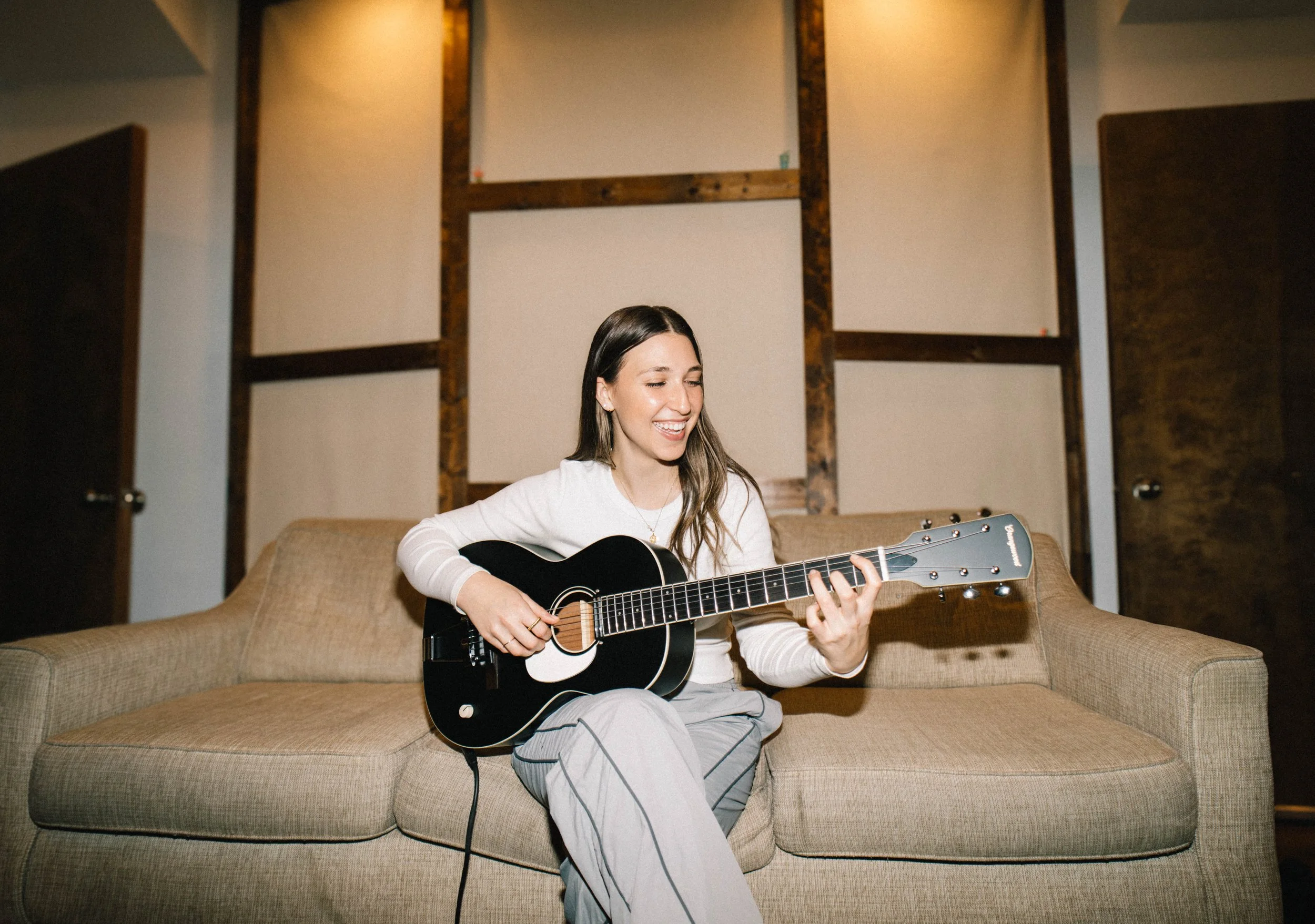 Julia DeTomaso smiling on beige couch with acoustic guitar — warm indoor studio portrait with wood-paneled walls.