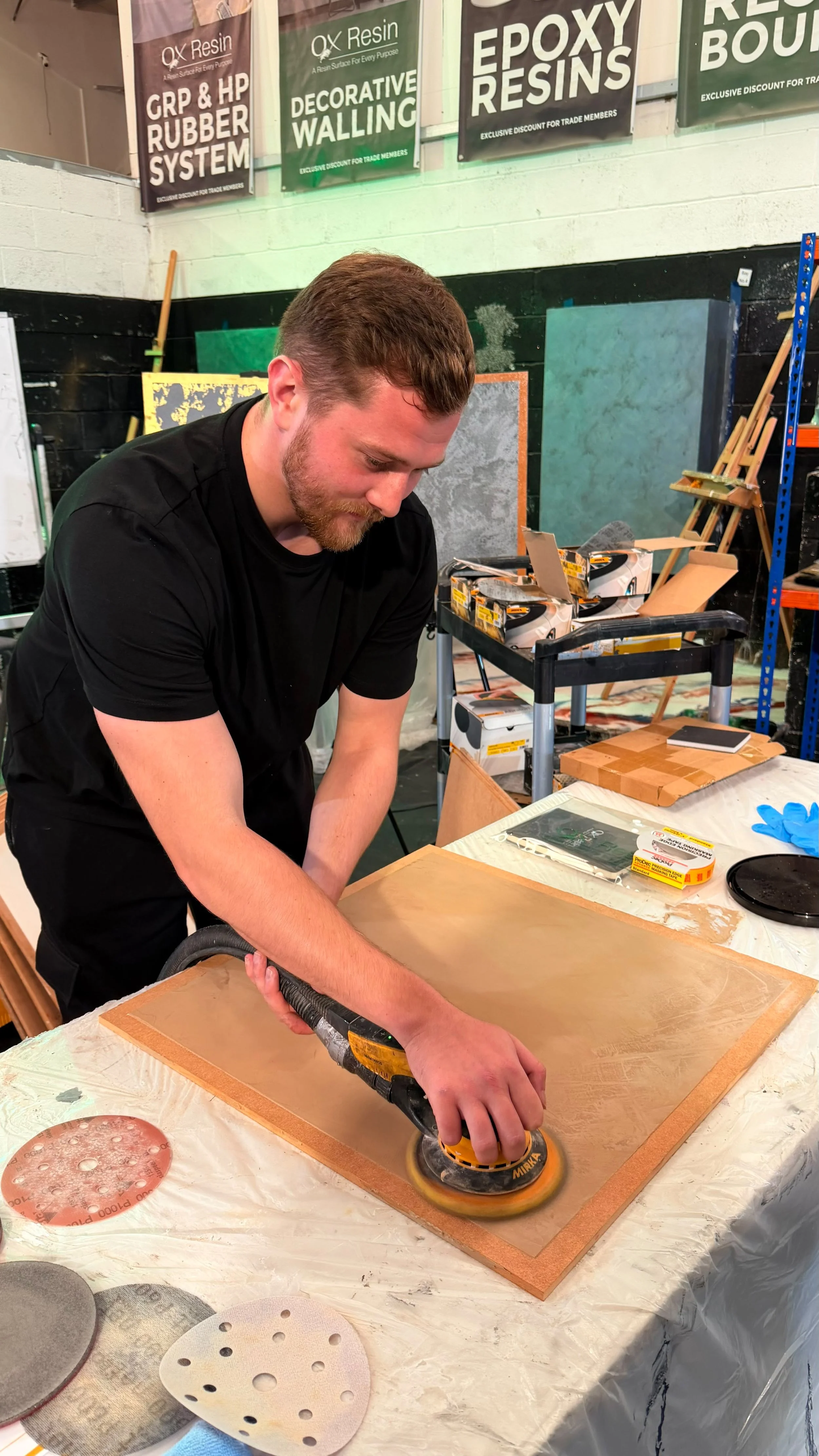 A man in a black T-shirt is polishing a wooden surface with an electric sander. There are sanding discs and related tools on the table. The background has industrial decor, with paint samples, banners about epoxy resin products, and equipment in a workshop setting.