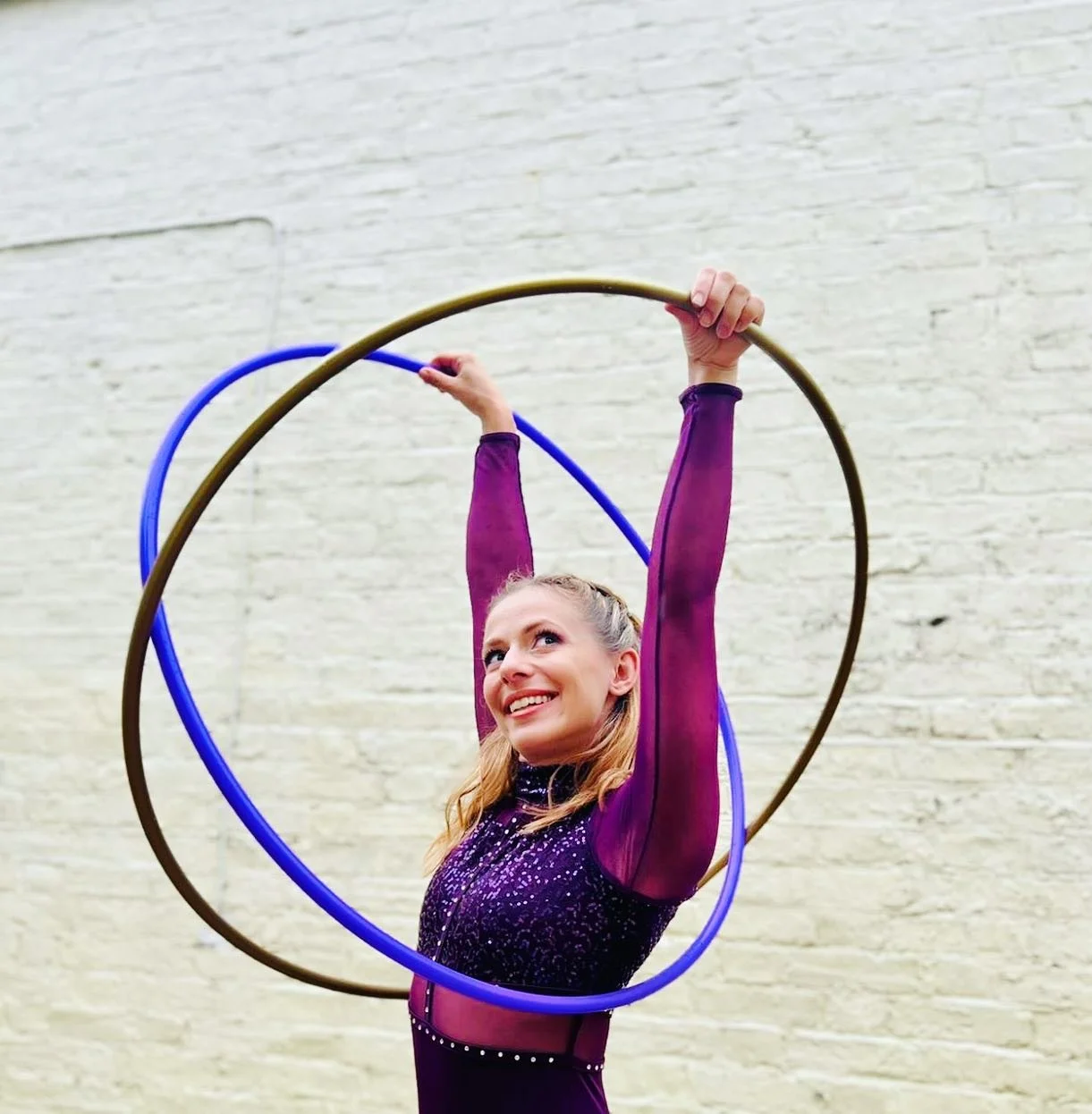 A female gymnast performing with two hula hoops, one gold and one blue, against a white brick wall, wearing a purple sparkly leotard with sheer purple sleeves, smiling and looking up.