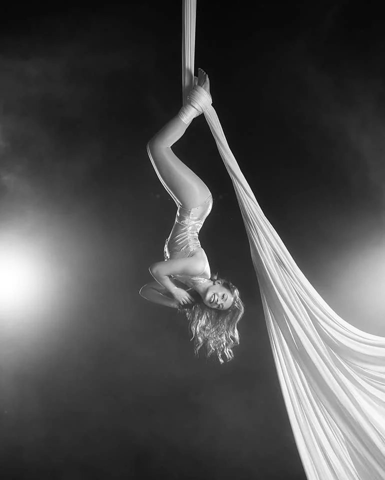 A female aerial silk performer hanging upside down on white silk fabric, smiling, in a black and white photo.
