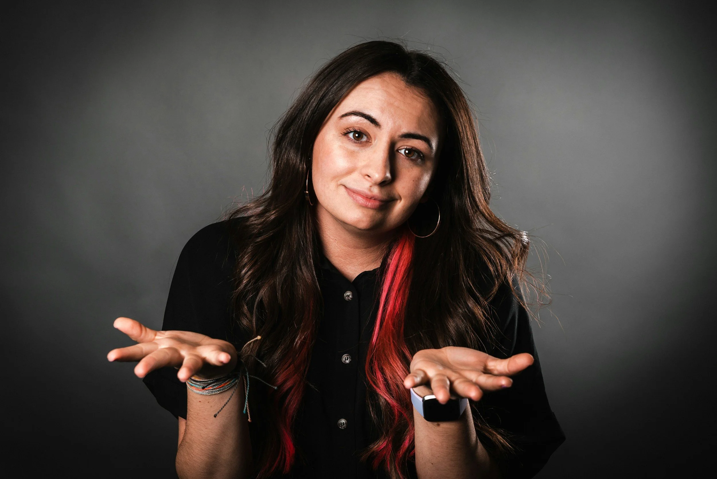 Young woman with long dark hair and red highlights, wearing a black shirt and hoop earrings, shrugging with a slight smile against a dark gray background.