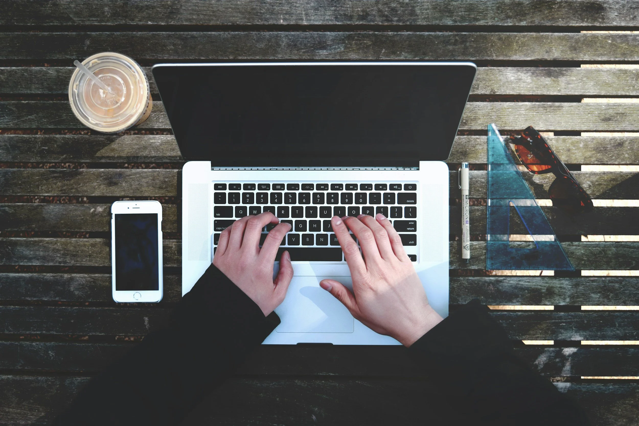 Top-down view of a person using a laptop on a wooden table, with a smartphone to the side, a coffee drink, sunglasses, a pen, and a transparent ruler nearby.