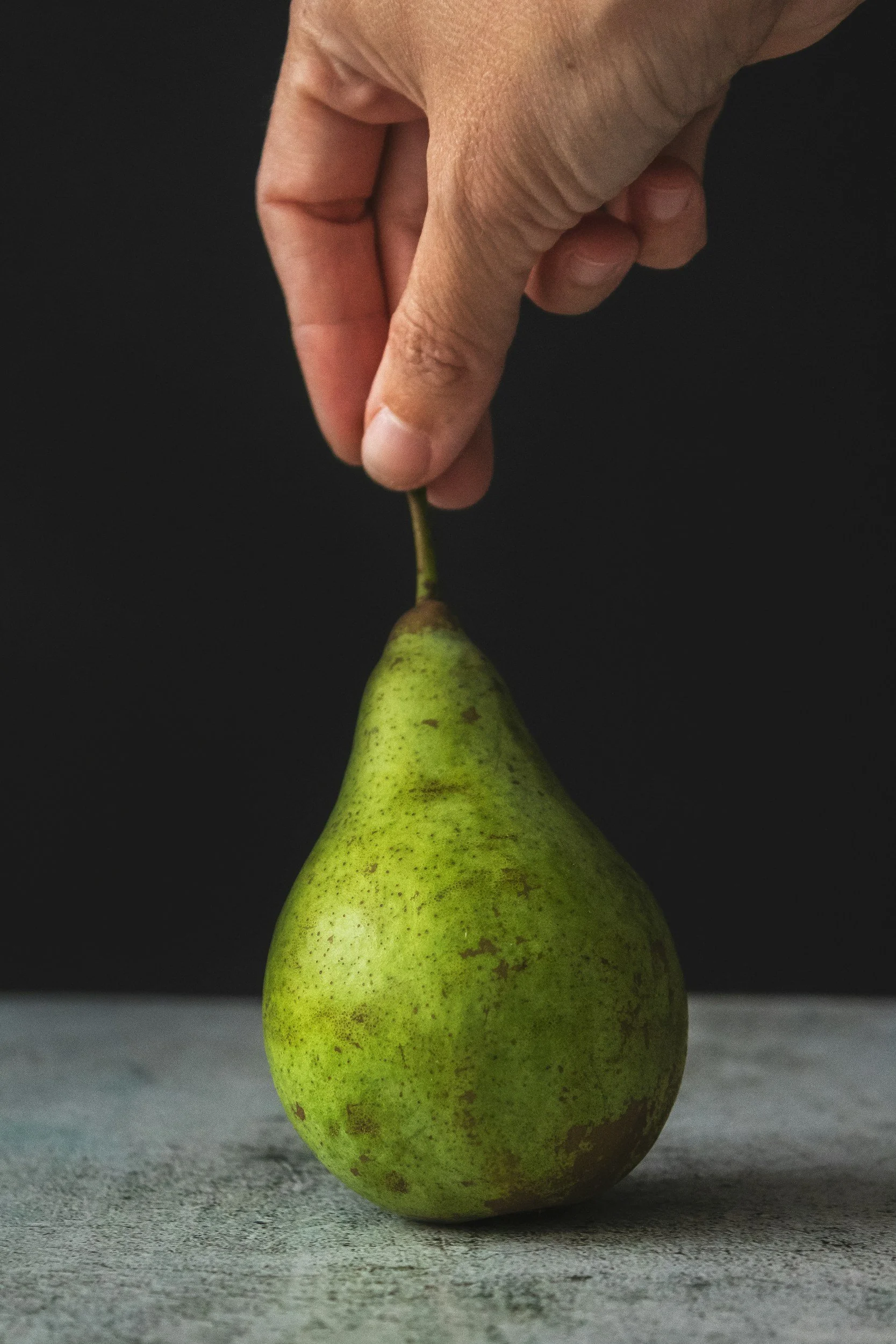 A close-up of a person's hand holding the stem of a green pear on a gray surface against a dark background.