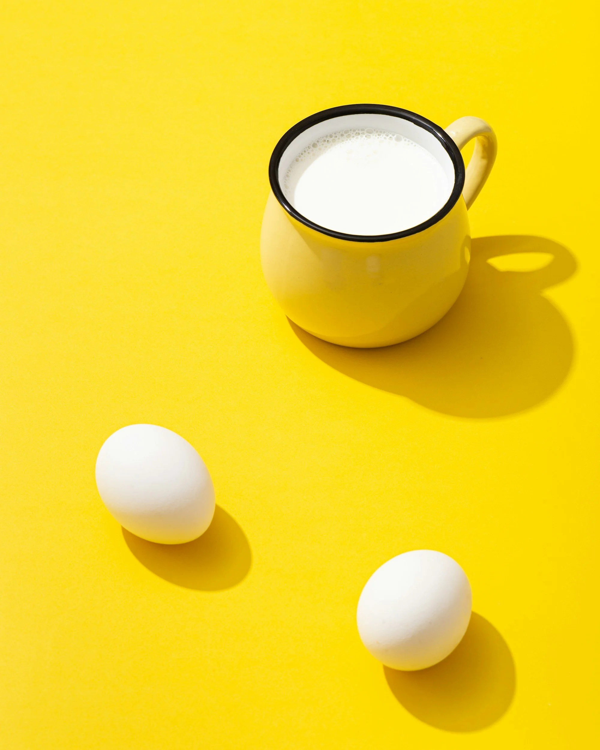 A yellow mug filled with milk, with two white eggs on a bright yellow surface, casting shadows.