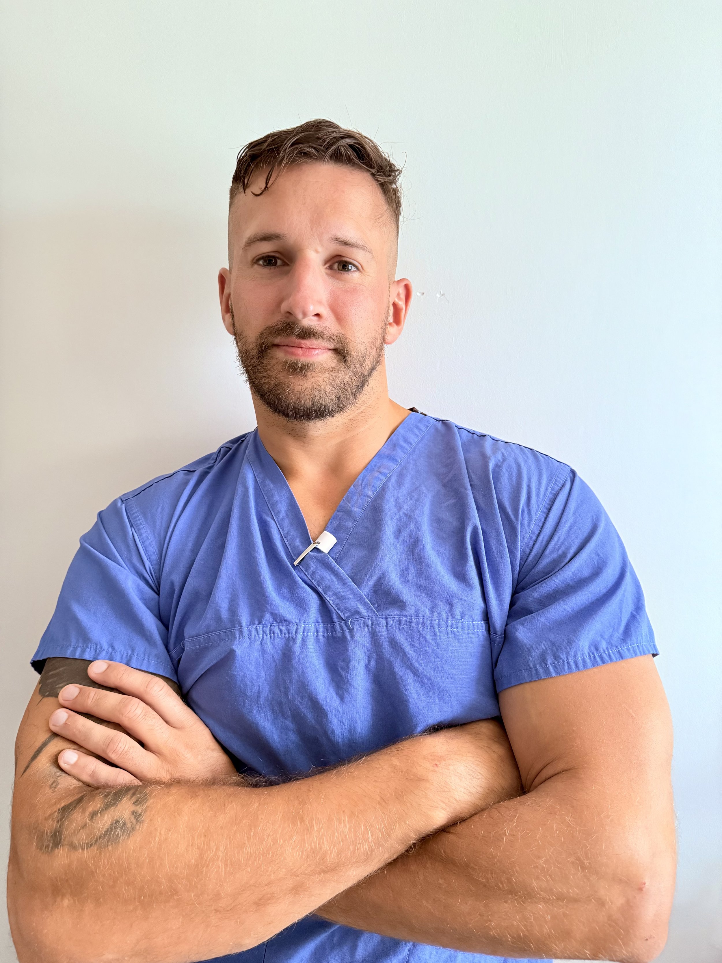 A male healthcare professional in blue scrubs standing against a light-colored wall with arms crossed.