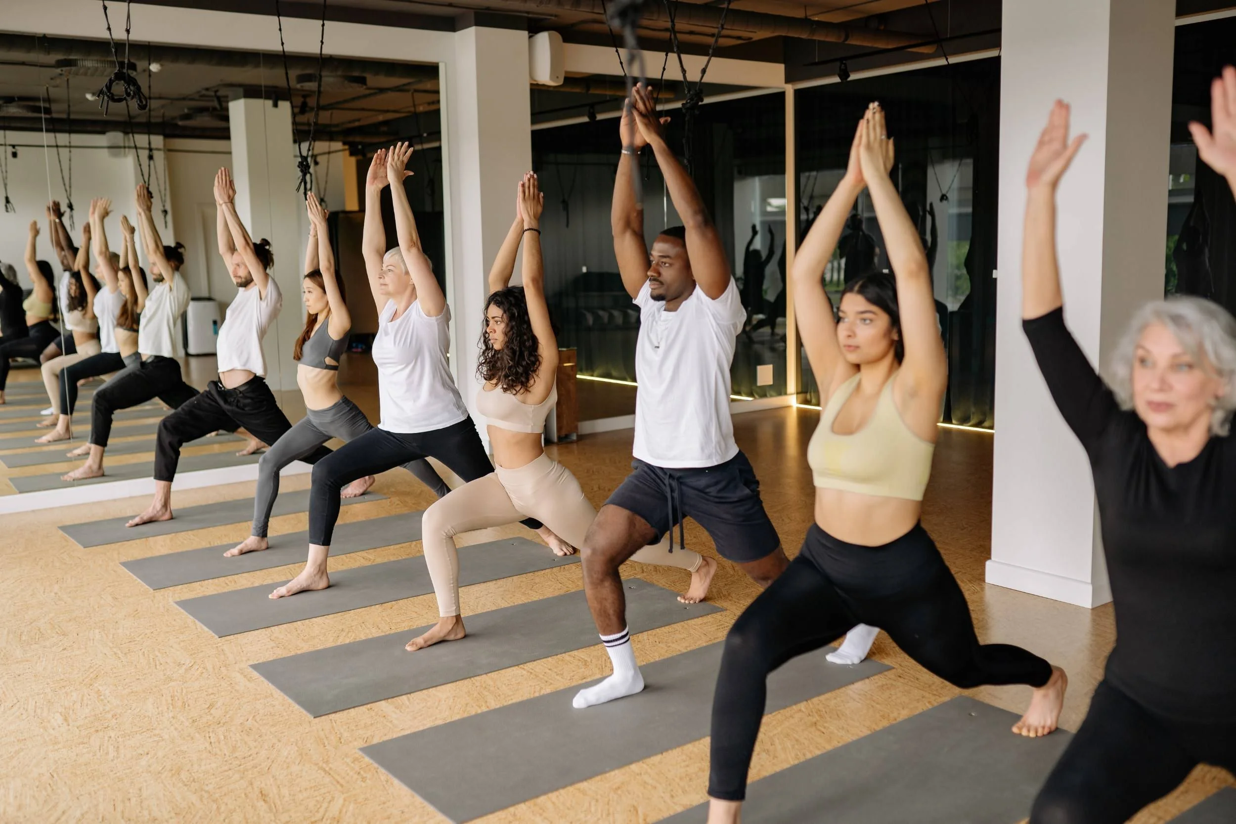 A group of diverse people in athletic wear practicing yoga in a studio, performing high mountain poses with their hands raised above their heads.
