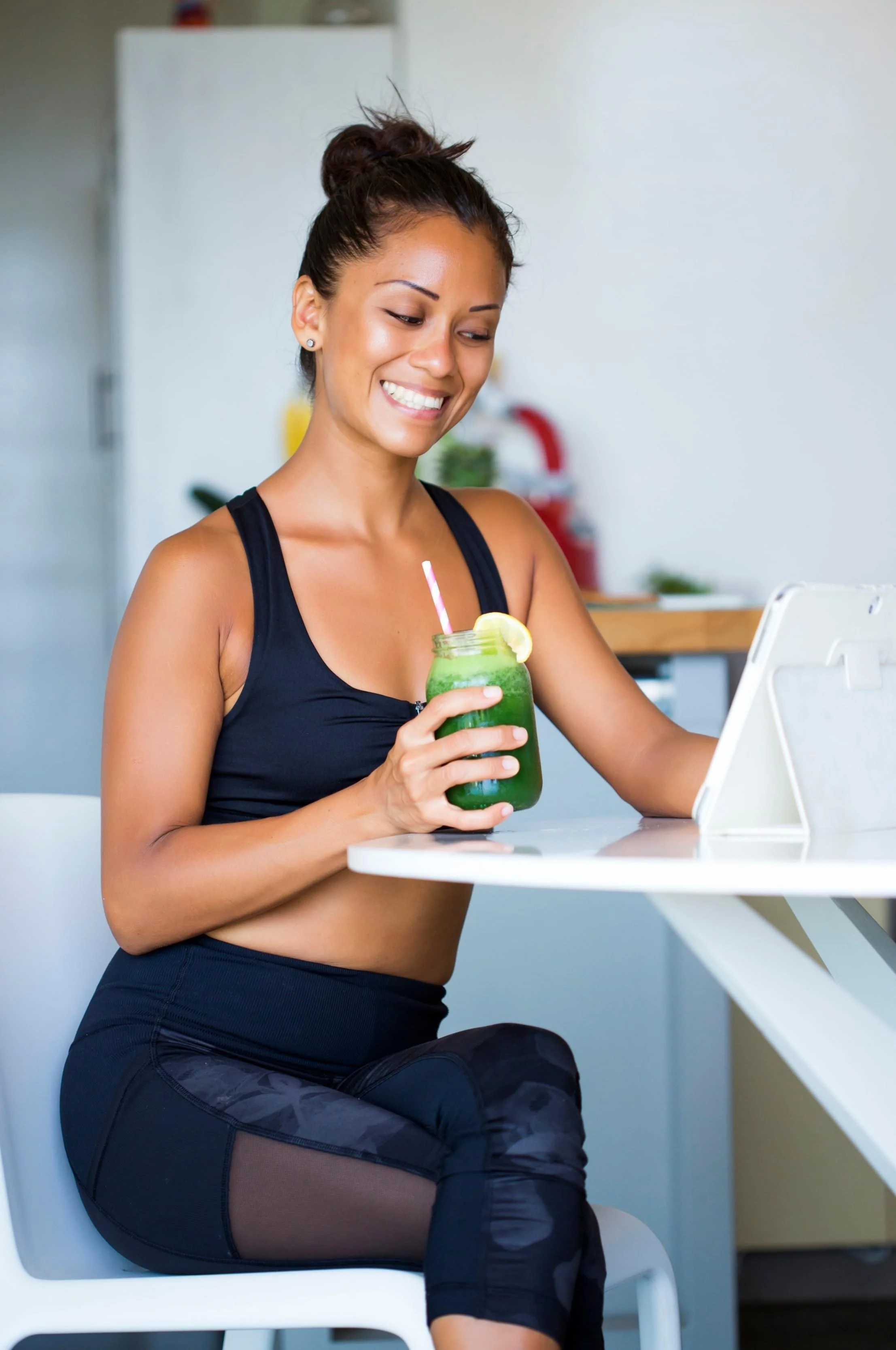 A woman in black workout clothing is smiling and sitting at a white table, holding a jar of green smoothie with a lemon slice and a pink and white striped straw, while looking at a tablet.
