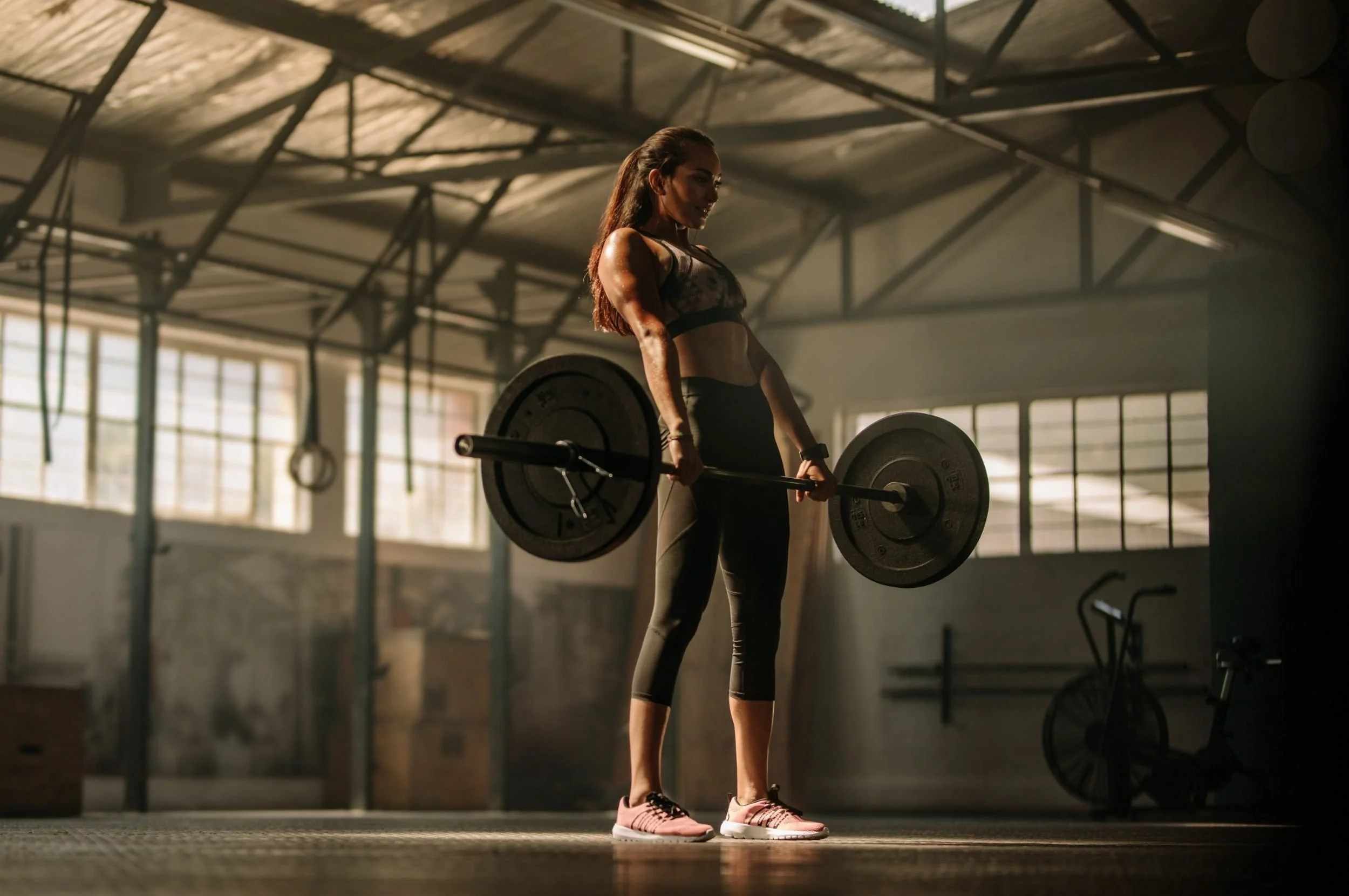 A woman with long hair in athletic wear and pink shoes lifting a barbell in a gym with large windows and high ceilings.