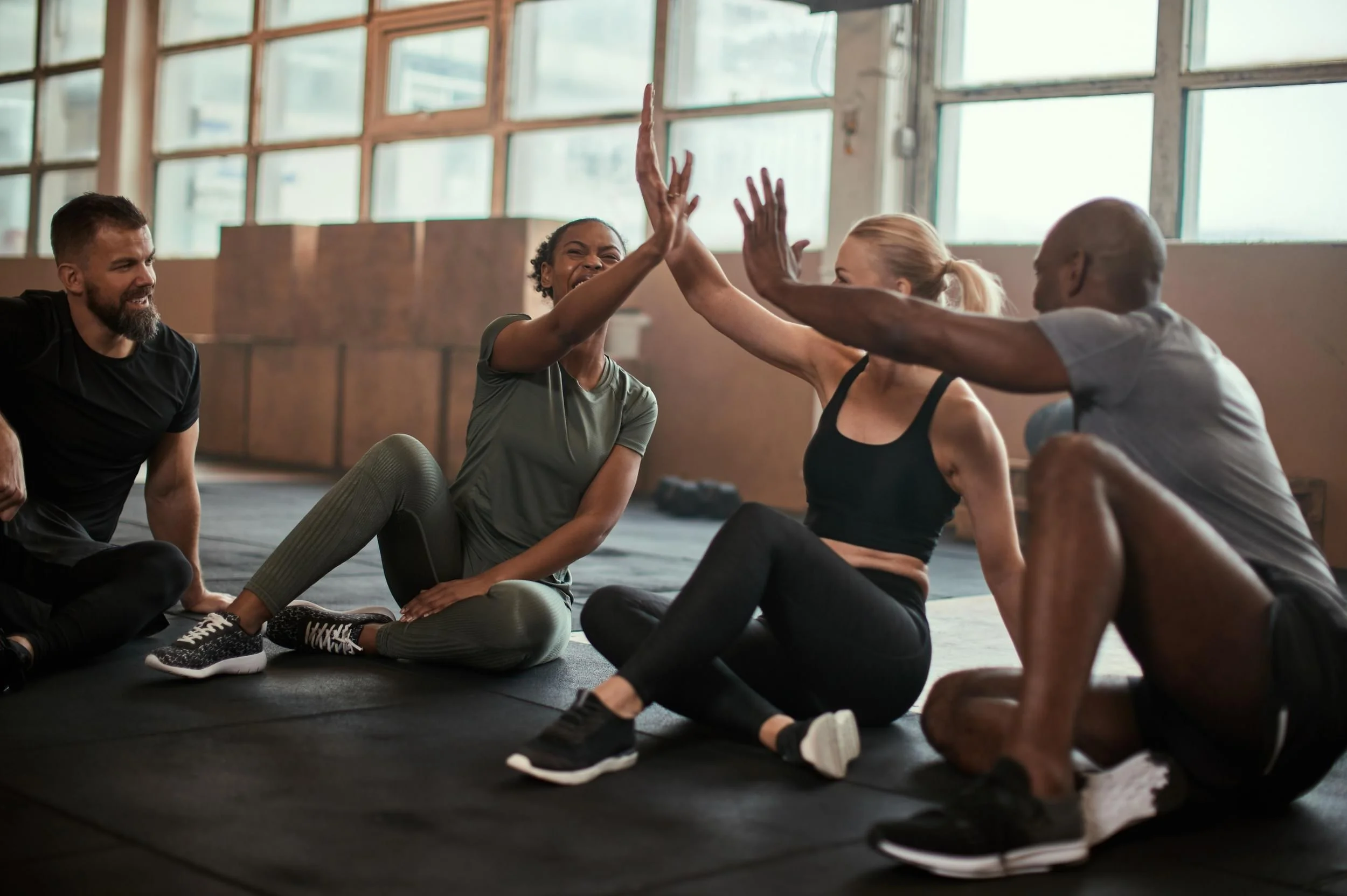 Four people sitting on the floor in a gym, engaging in a friendly high-five. They are casually dressed, with gym equipment and large windows in the background.